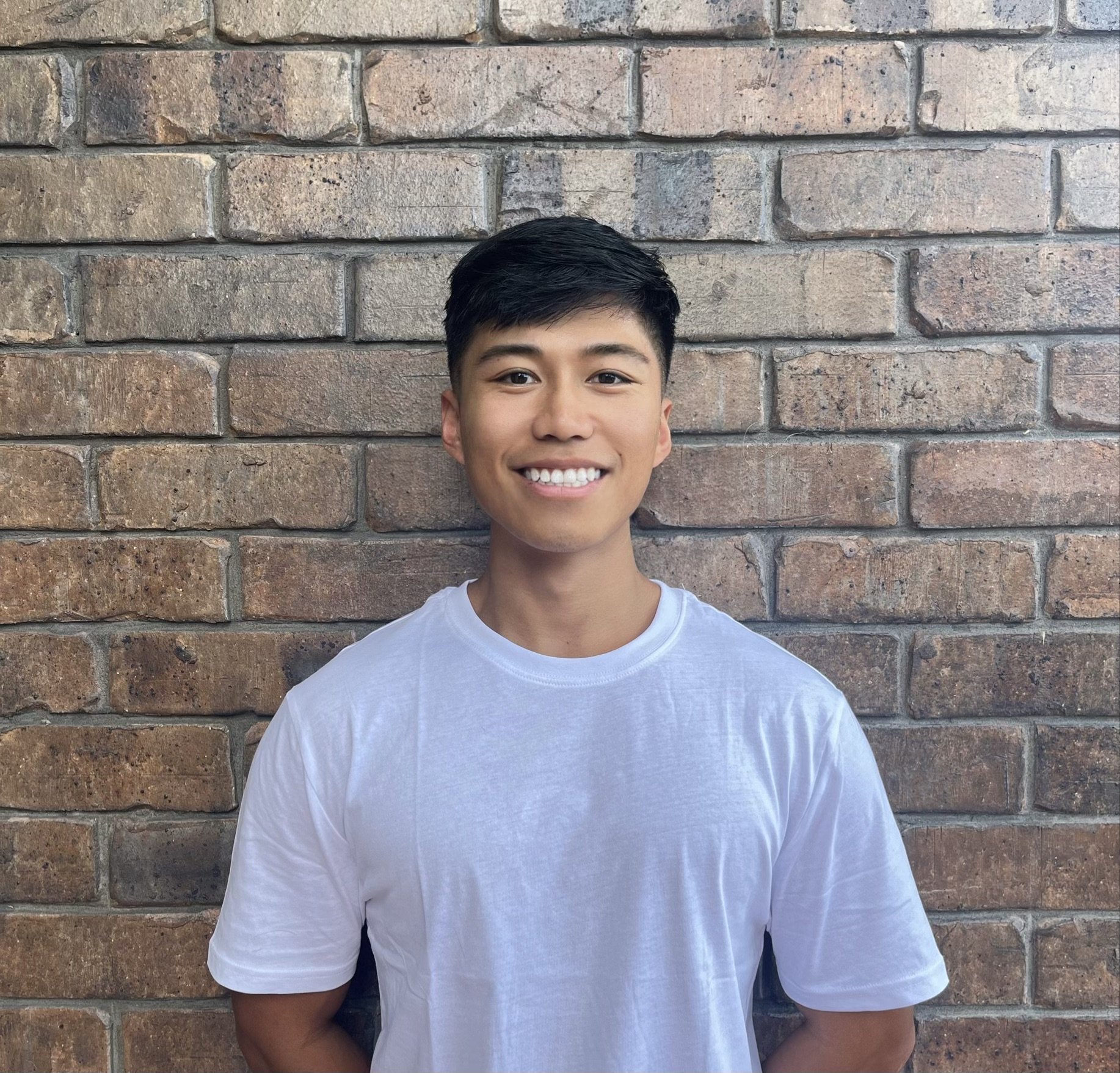 Young man with dark hair smiling in front of a brick wall, wearing a white T-shirt.