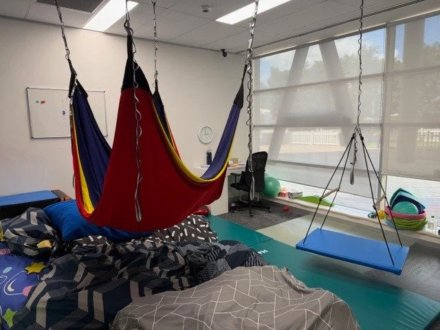 Indoor physical therapy or exercise room with a bed, hanging hammock, a swing, exercise balls, and various fitness equipment, with large windows and blinds.