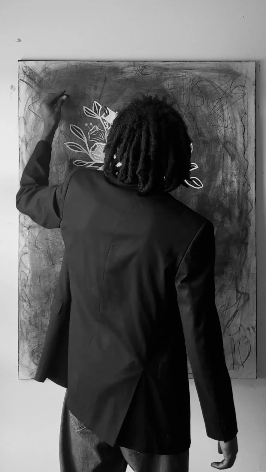 Black-and-white photo of a person in a blazer drawing leafy botanical designs on a large wall-mounted canvas.