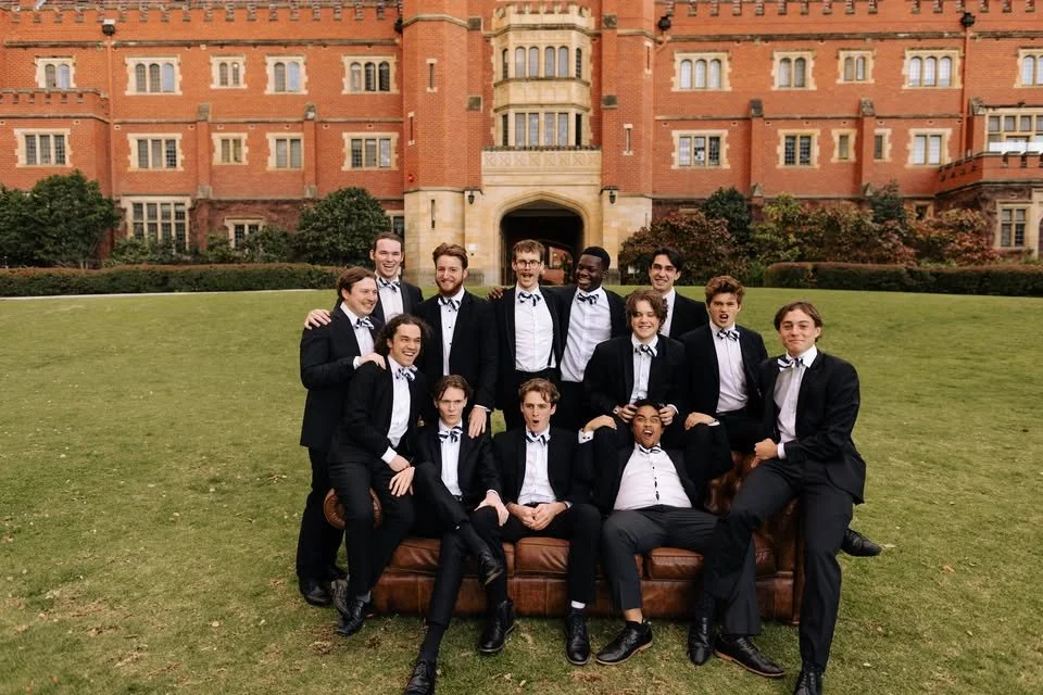 13 young men sit on a brown leather counch on the front lawn of St George's College.