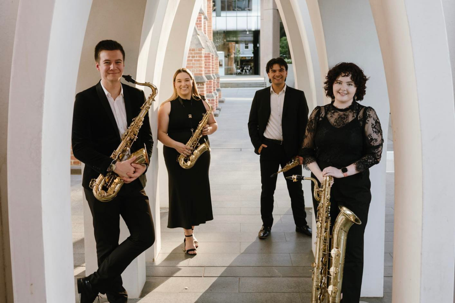 Blake Bayley, Grace Kay, Joshua Sumich and Andrea Jordan-Keane holding saxophones, standing under white arches outdoors, dressed in black formal attire, smiling, with a building and street in the background.