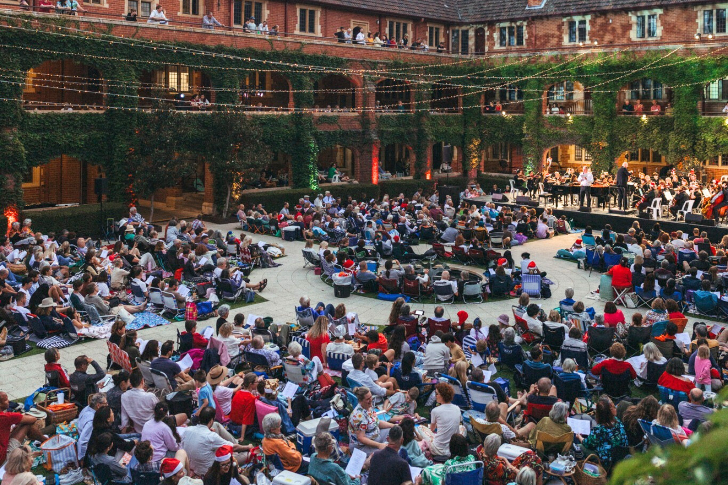 An outdoor concert at St George's with many people seated in chairs and on blankets, watching a musical performance on stage. The courtyard is decorated with string lights and surrounded by a brick building covered in green vines.