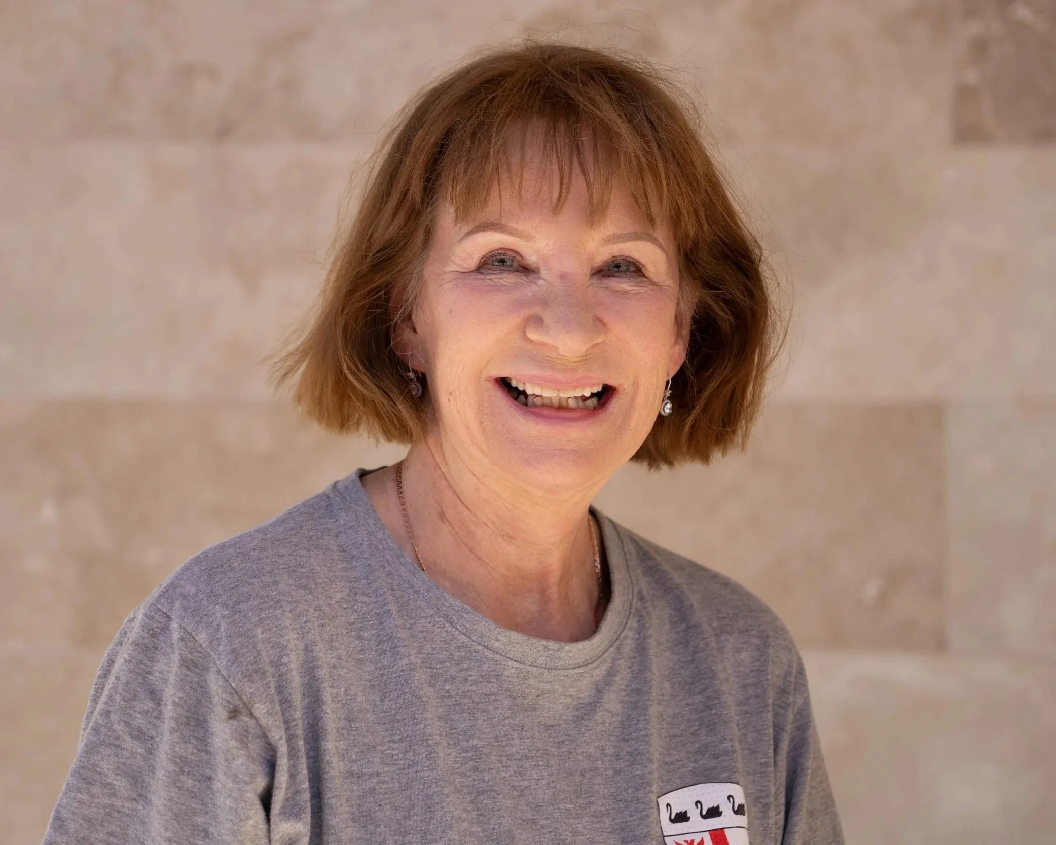 A smiling middle-aged woman with short red hair, wearing a gray T-shirt and earrings, standing in front of a beige tiled wall.