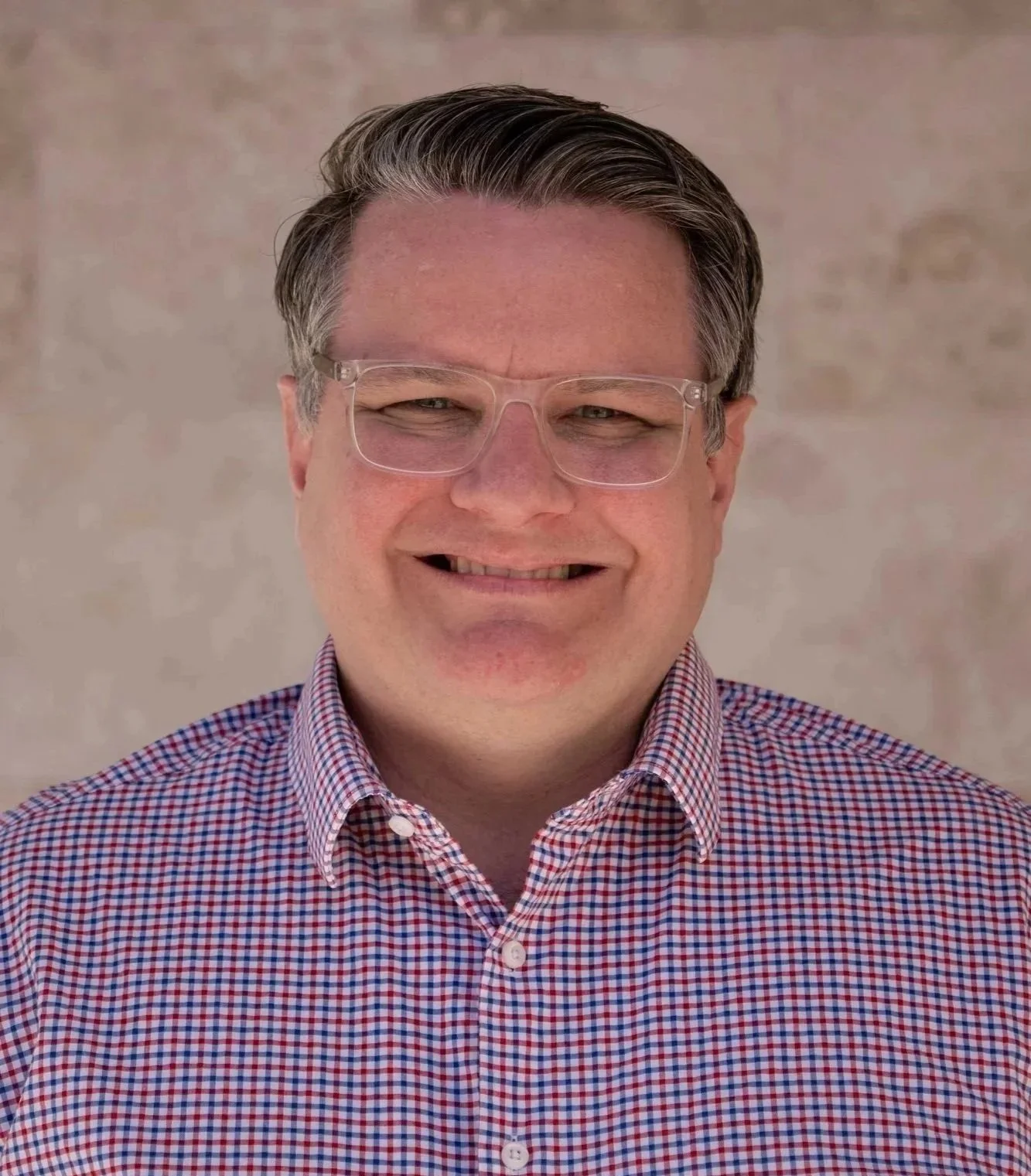 A smiling man with glasses and short dark hair wearing a red and white checkered shirt.