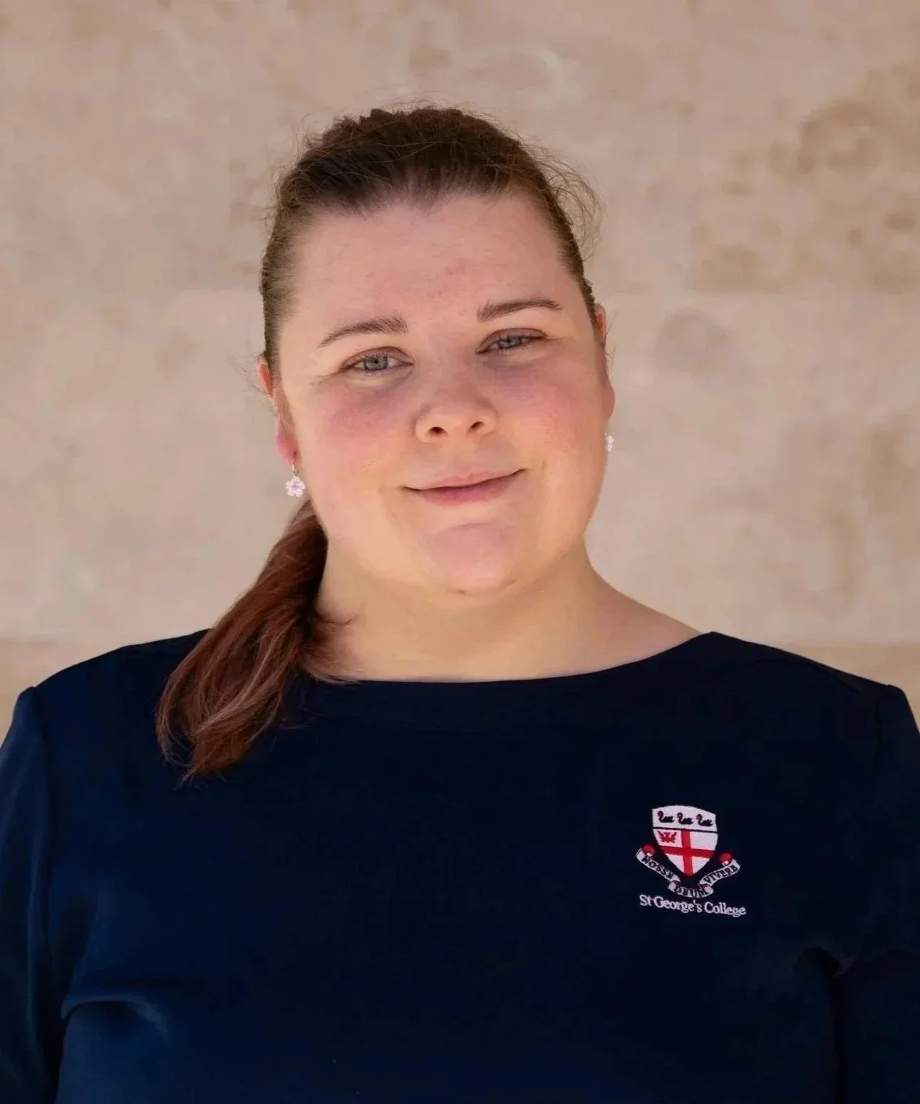 A woman with brown hair pulled back, wearing earrings, and a navy blue shirt with a St. George's College logo.