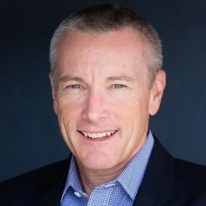 A middle-aged man with short gray hair, wearing a blue checkered shirt and a dark blazer, smiling against a dark background.