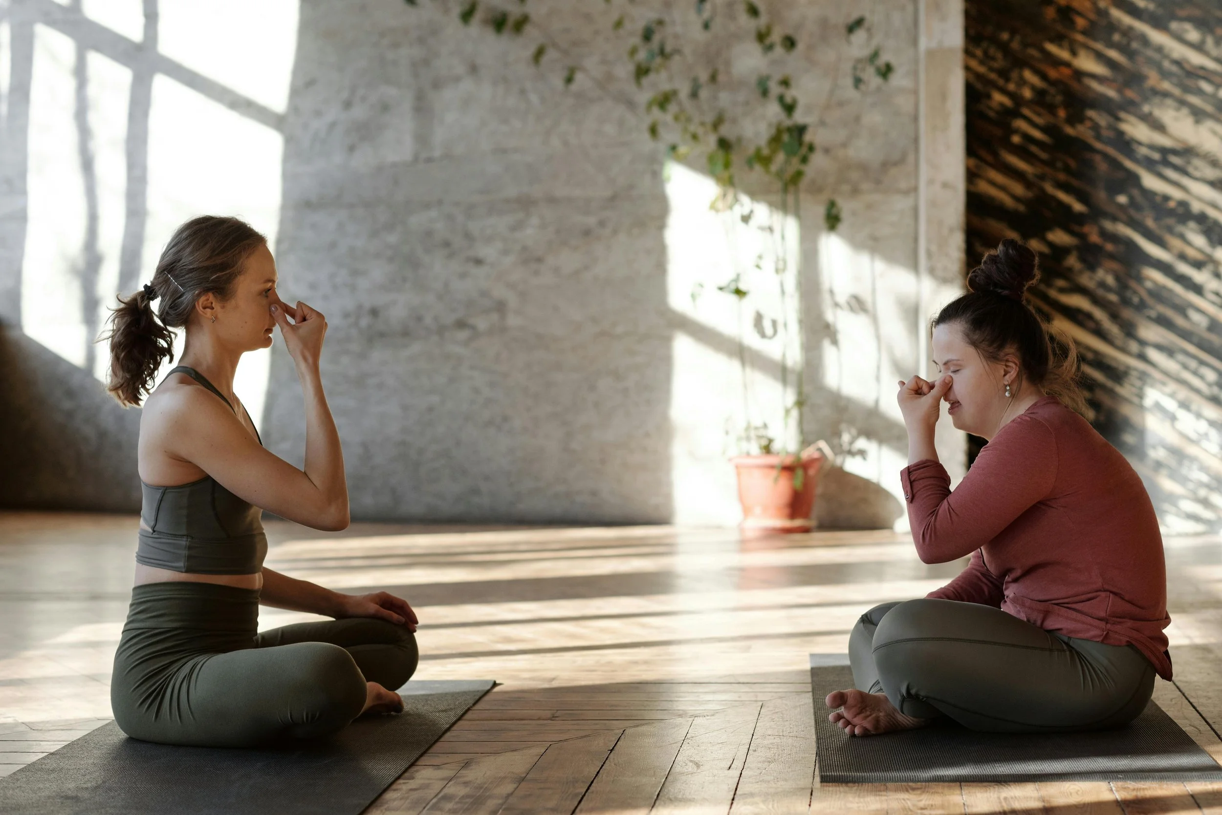 Twee vrouwen doen ademtechnieken -de wisselende neusgat ademhaling- in een open lichte ruimte met houten vloer, een plan achteraan. Ze zitten op twee grijze yogamatten en eentje heeft een bordeaux outfit.