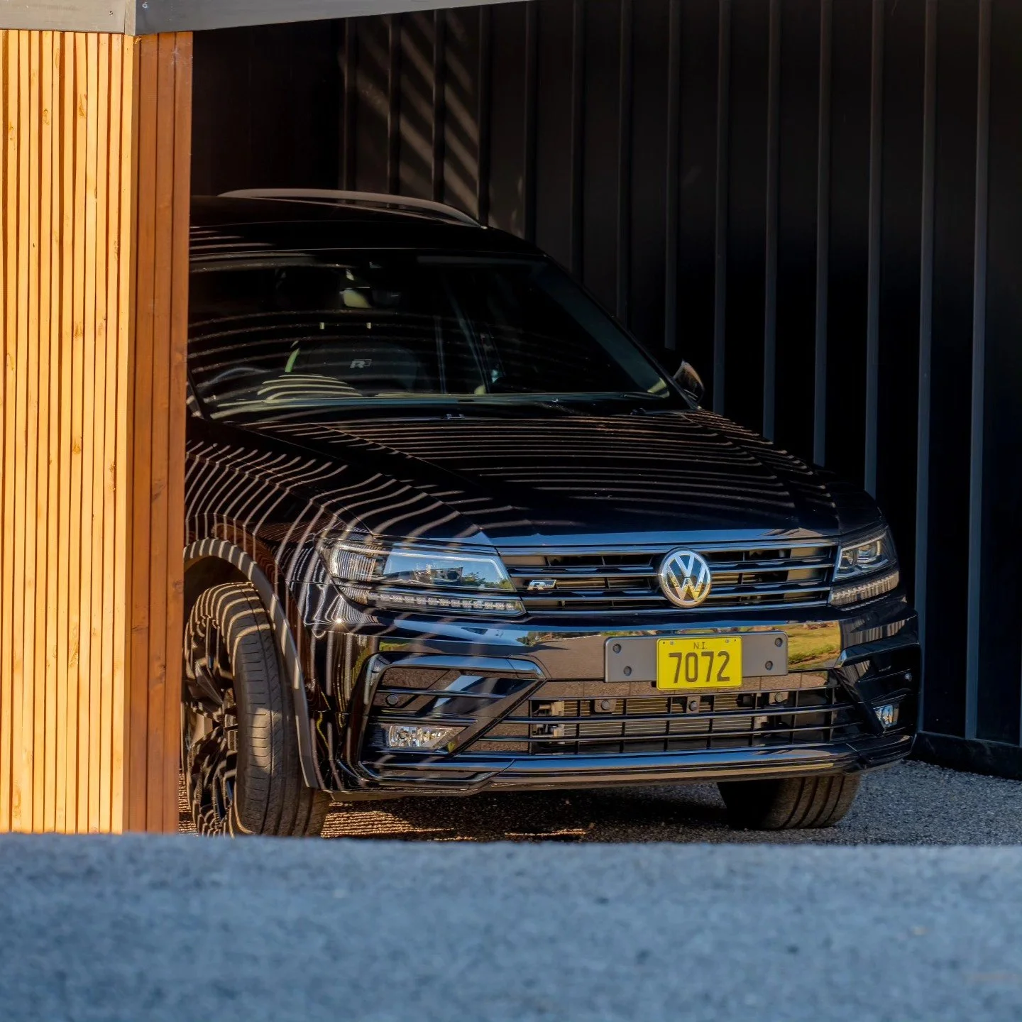 A black Volkswagen car parked in a driveway, partially obscured by a wooden fence on the left and a dark fence on the right side.