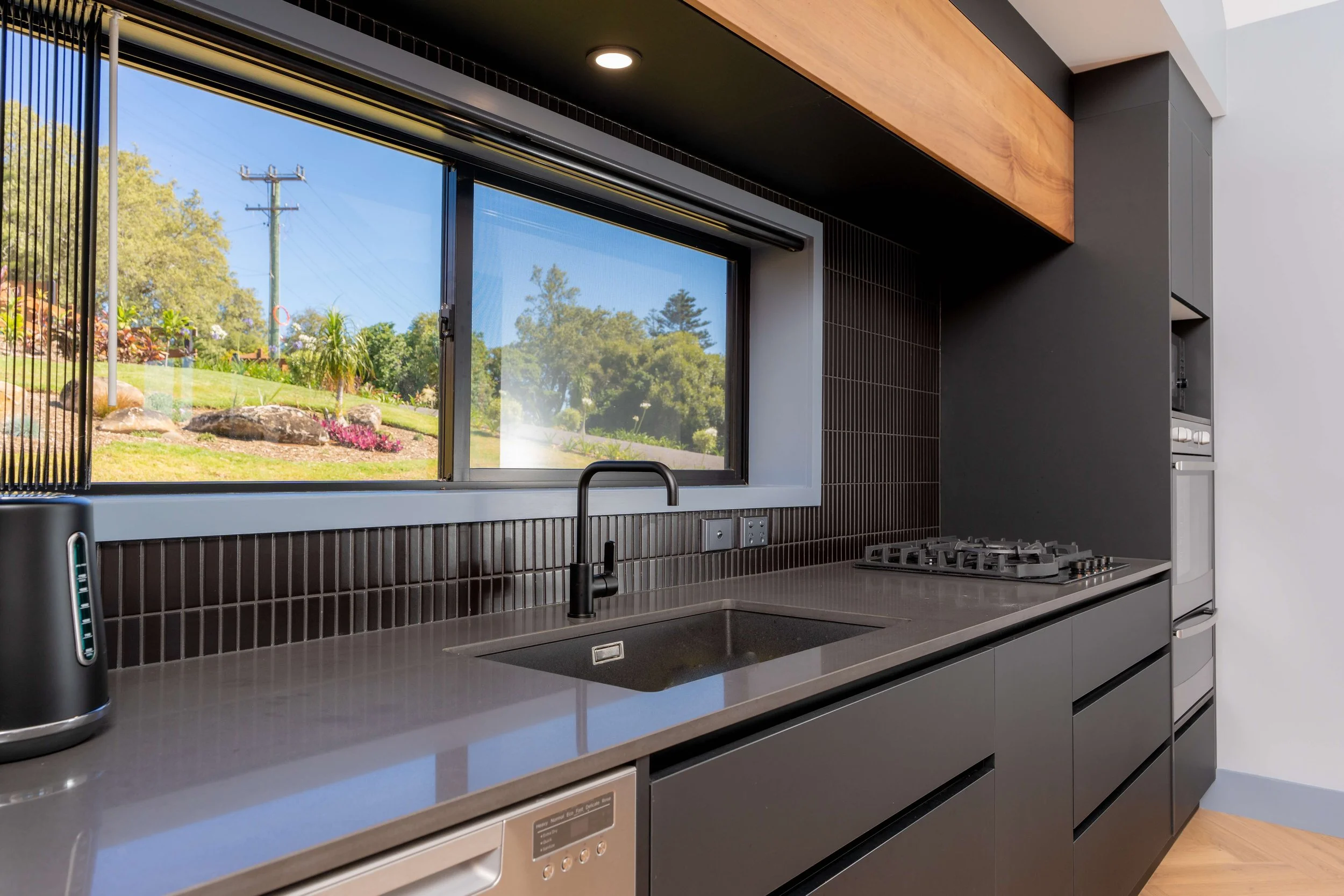 Modern kitchen with a large window overlooking a garden, black backsplash, gray countertop, and built-in appliances.