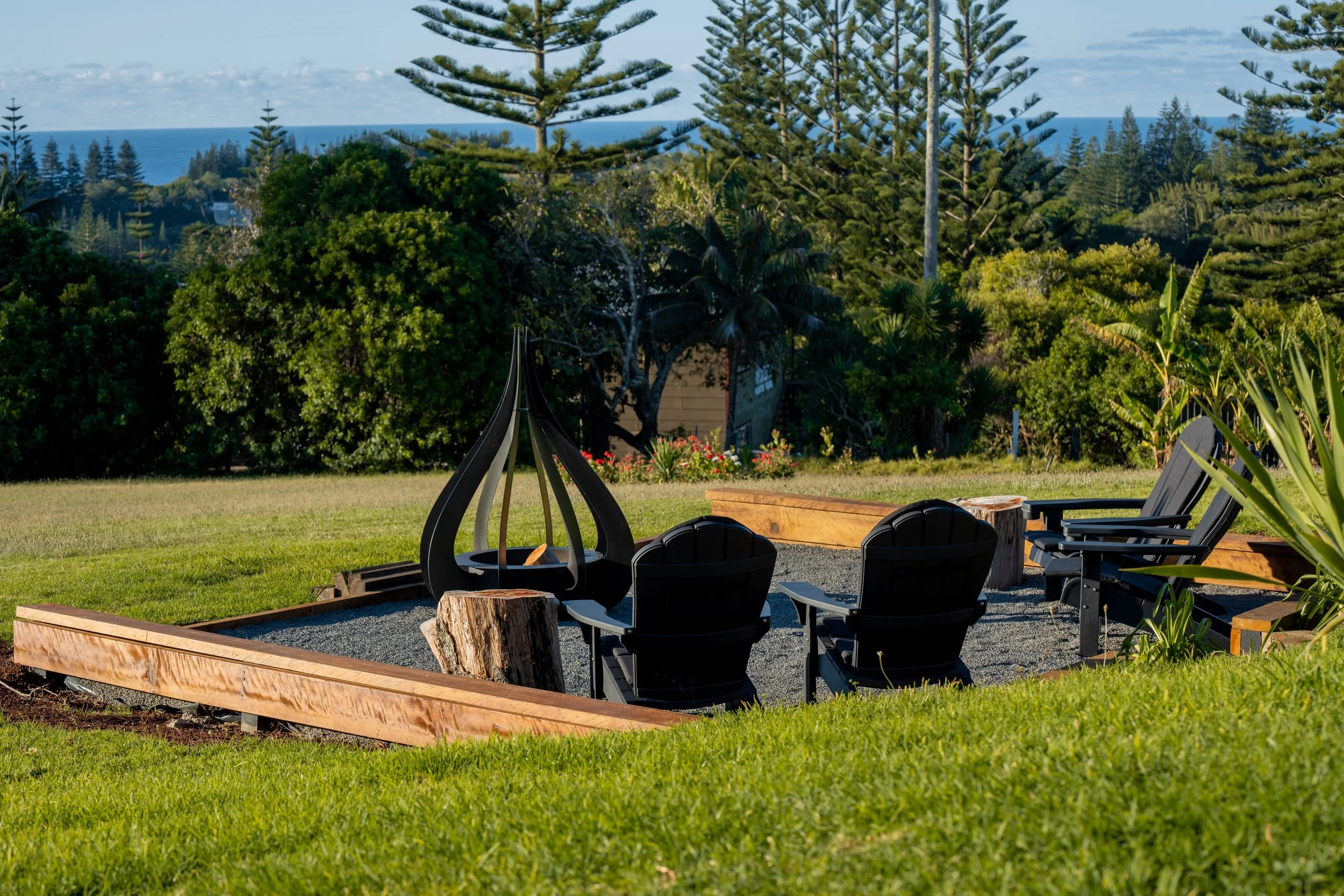 Outdoor fire pit seating area with black chairs, a metal fire bowl, and a tree stump for a table, surrounded by lush greenery and overlooking the ocean in the background.