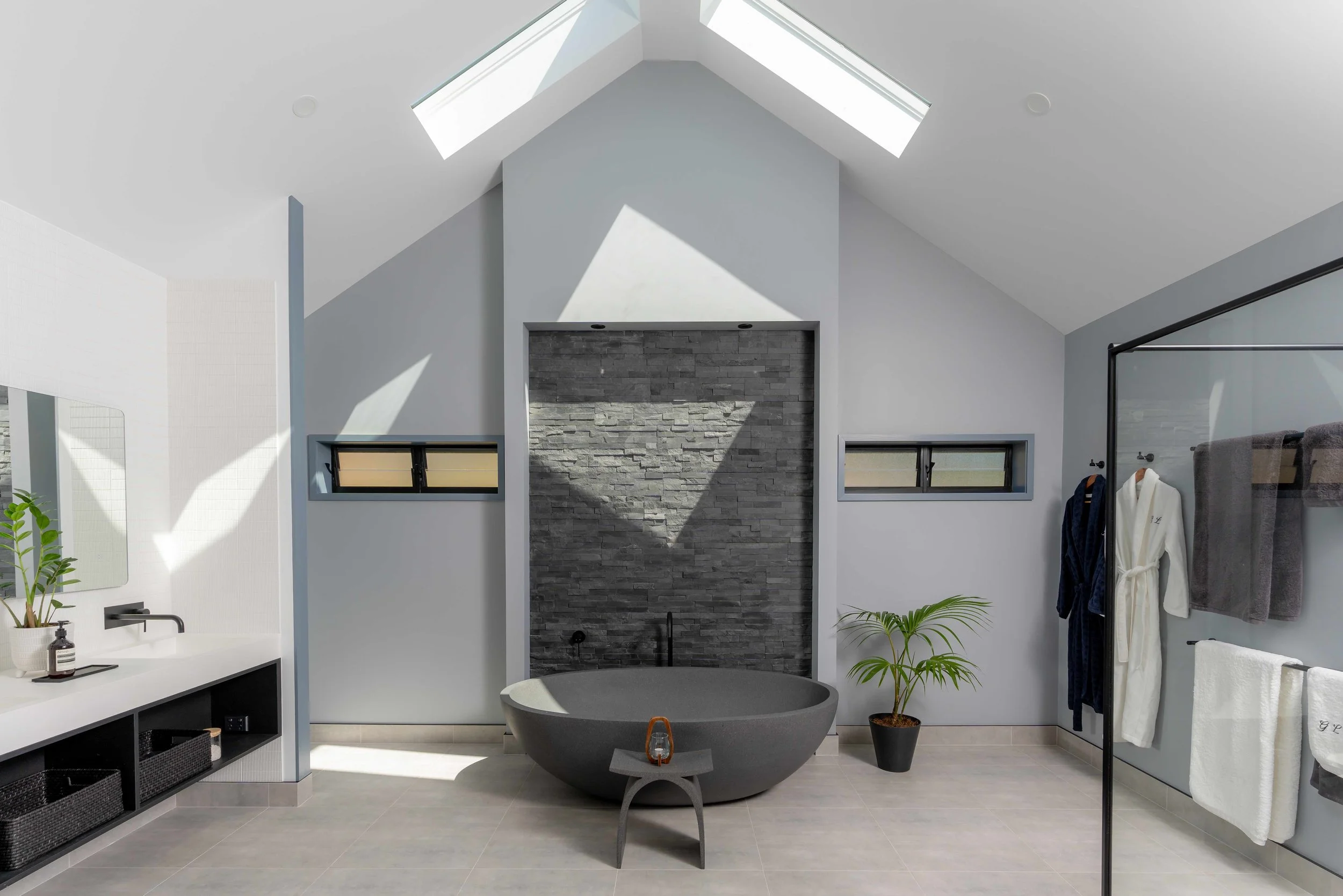 Modern bathroom featuring a freestanding round gray bathtub, small gray stool with a lantern, black towel rack with white and gray towels, a white robe hanging, a black potted plant, and a stone accent wall, with skylights above bringing natural ligh