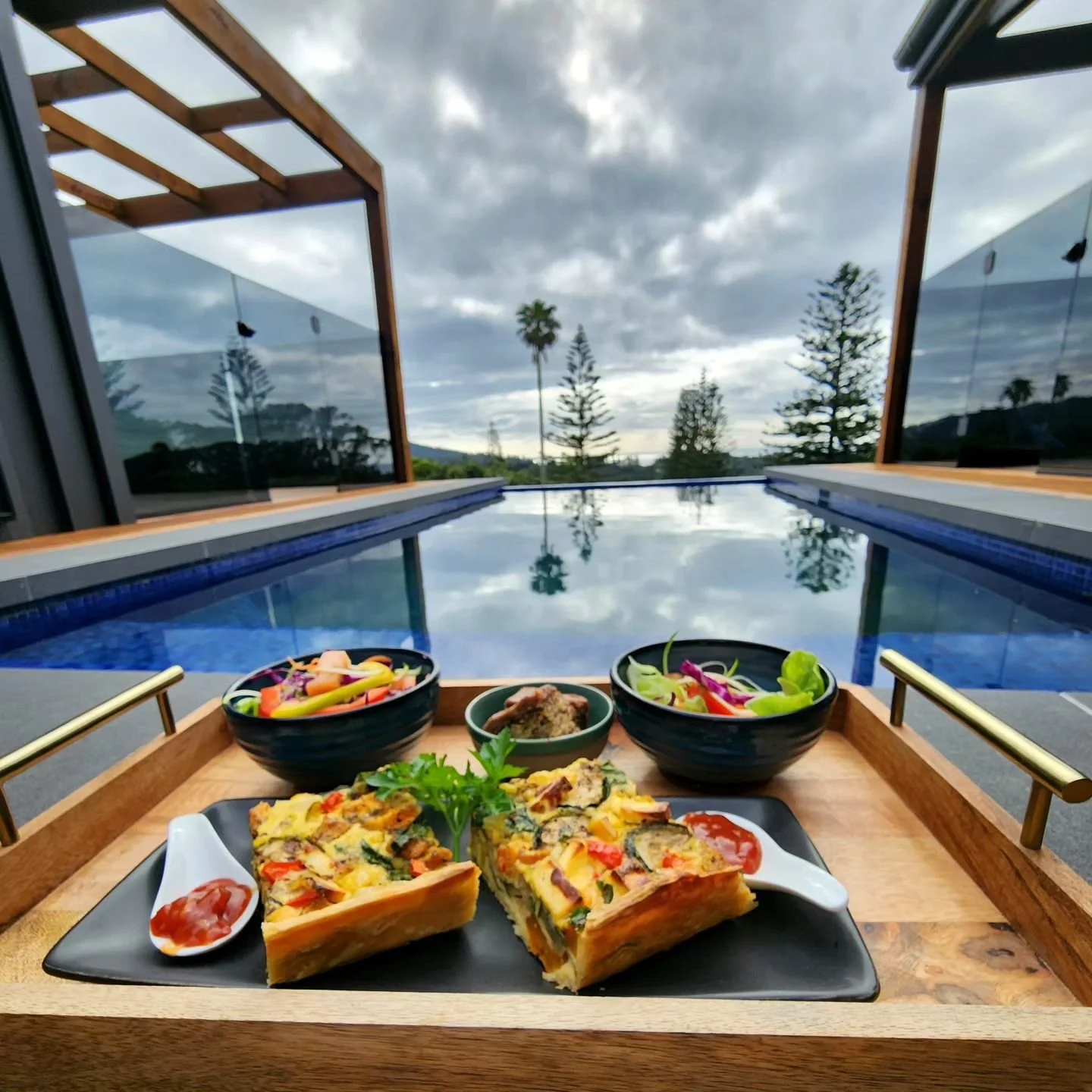 A tray with two slices of vegetable quiche, two small bowls of salad, and two bowls of additional salad dishes, set against a poolside outdoor background.