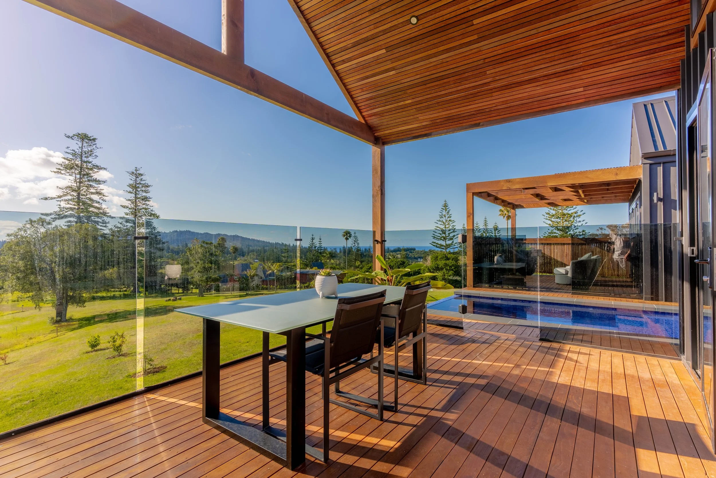 Outdoor patio with wooden flooring, glass railing, and a rectangular table with chairs, overlooking a green landscape with trees and hills under a clear blue sky.