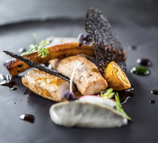 A gourmet dish with a piece of cooked fish, roasted vegetables, edible flowers, garnishes, and decorative sauces on a black plate.