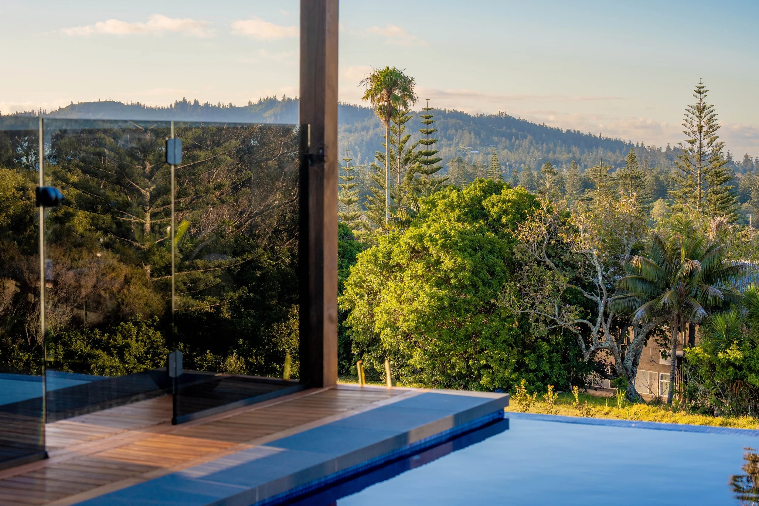 An outdoor scene with a glass enclosure and wooden deck overlooking lush green trees and a distant mountain range under a partly cloudy sky.