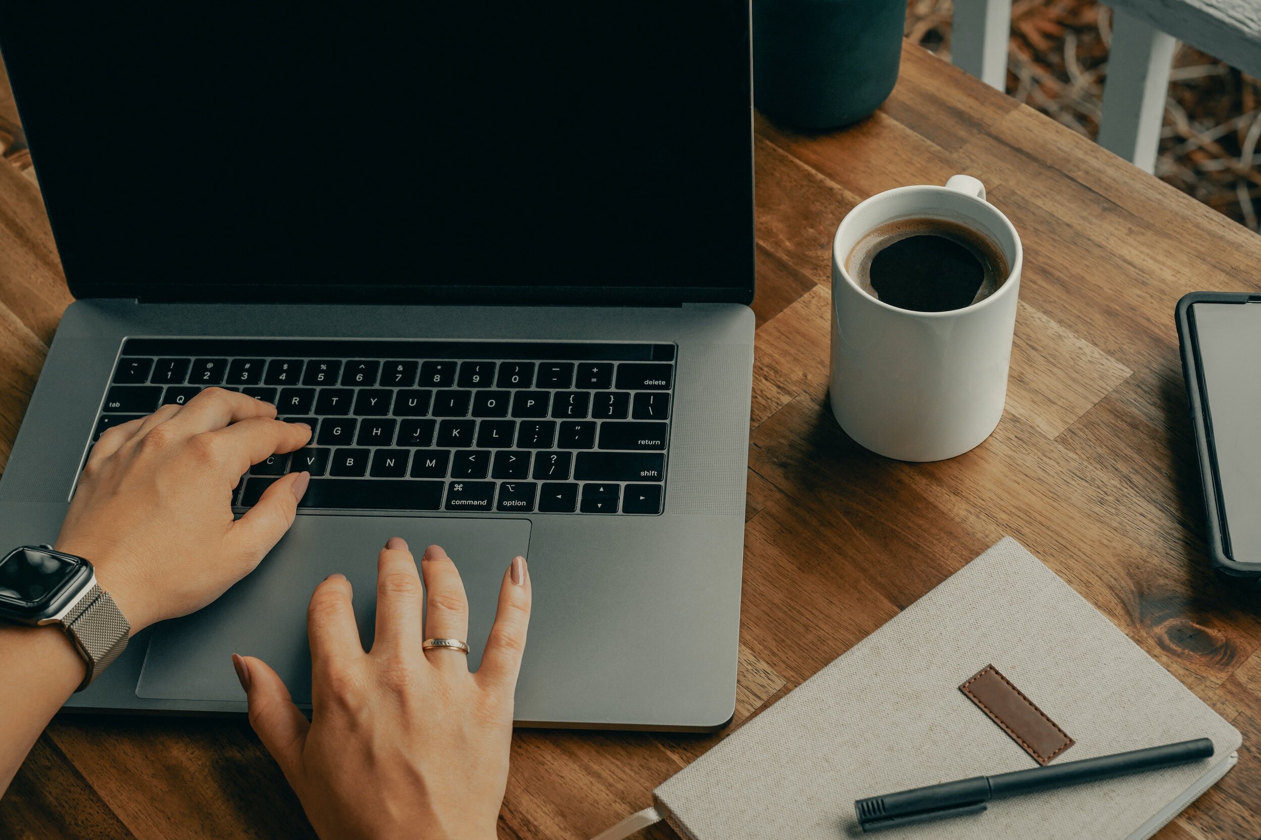 Virtual Assistant working on macbook with cup of black coffee in white mug and linen notebook with black pen on wooden table.