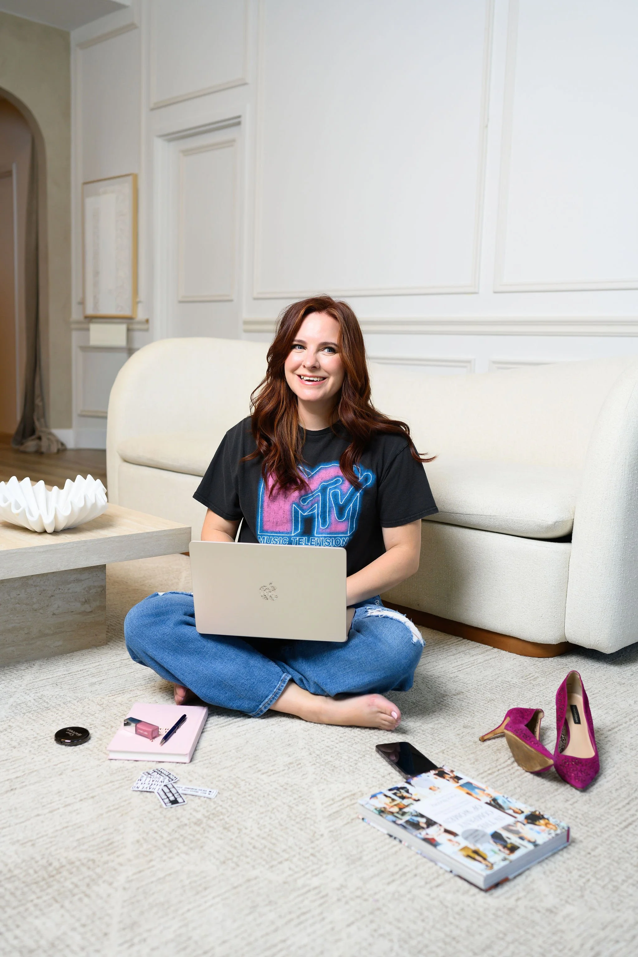 A woman with red hair sits on the floor in a living room, smiling at a laptop. She is wearing a black MTV T-shirt and ripped jeans. In front of her are makeup items, a smartphone, and magazines on a cream-colored carpet. Pink high heels are next to her on the right.