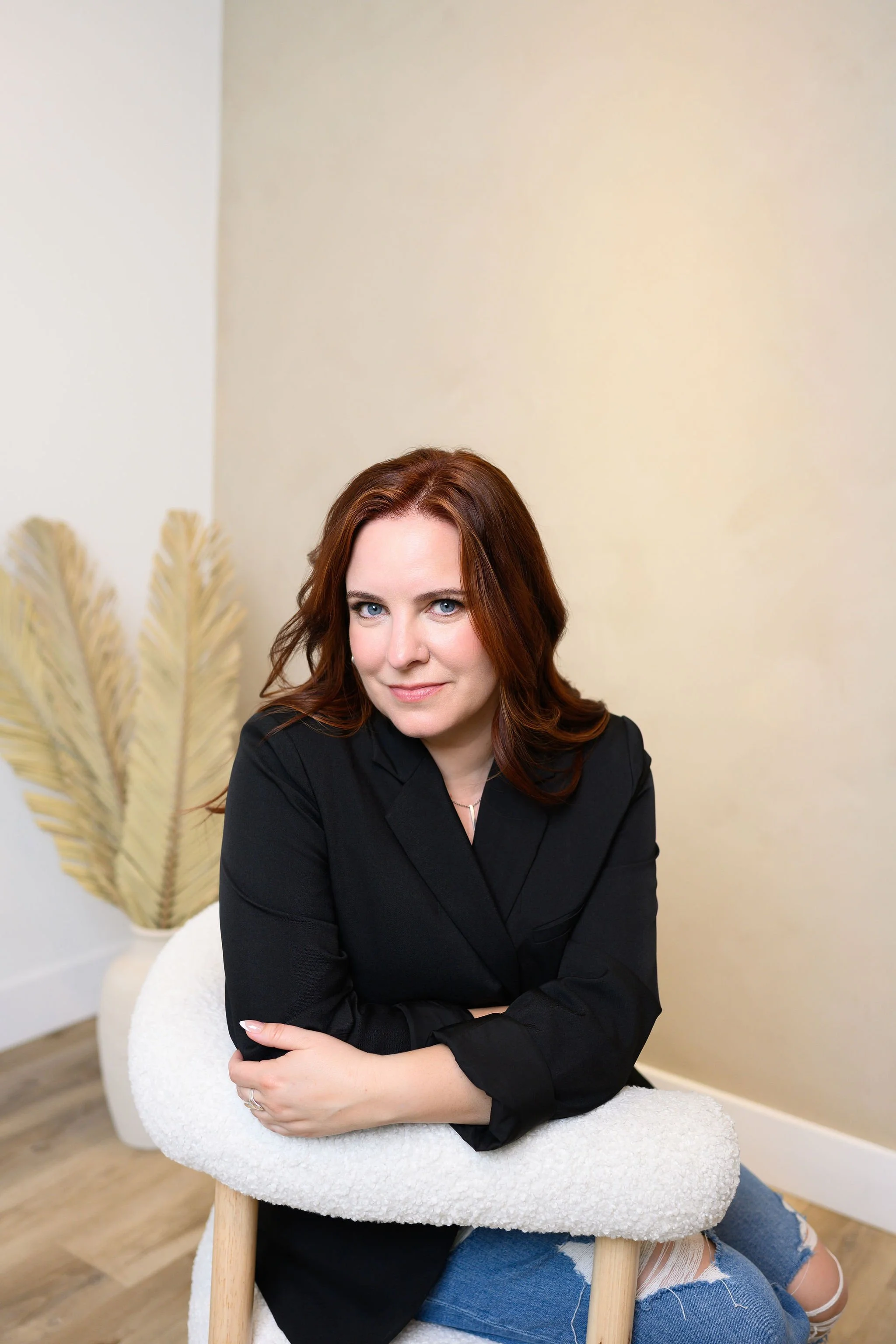 A woman with red hair and blue eyes sitting on a white chair, leaning on a fluffy white cushion, in a room with cream-colored walls and dried pampas grass in the background.