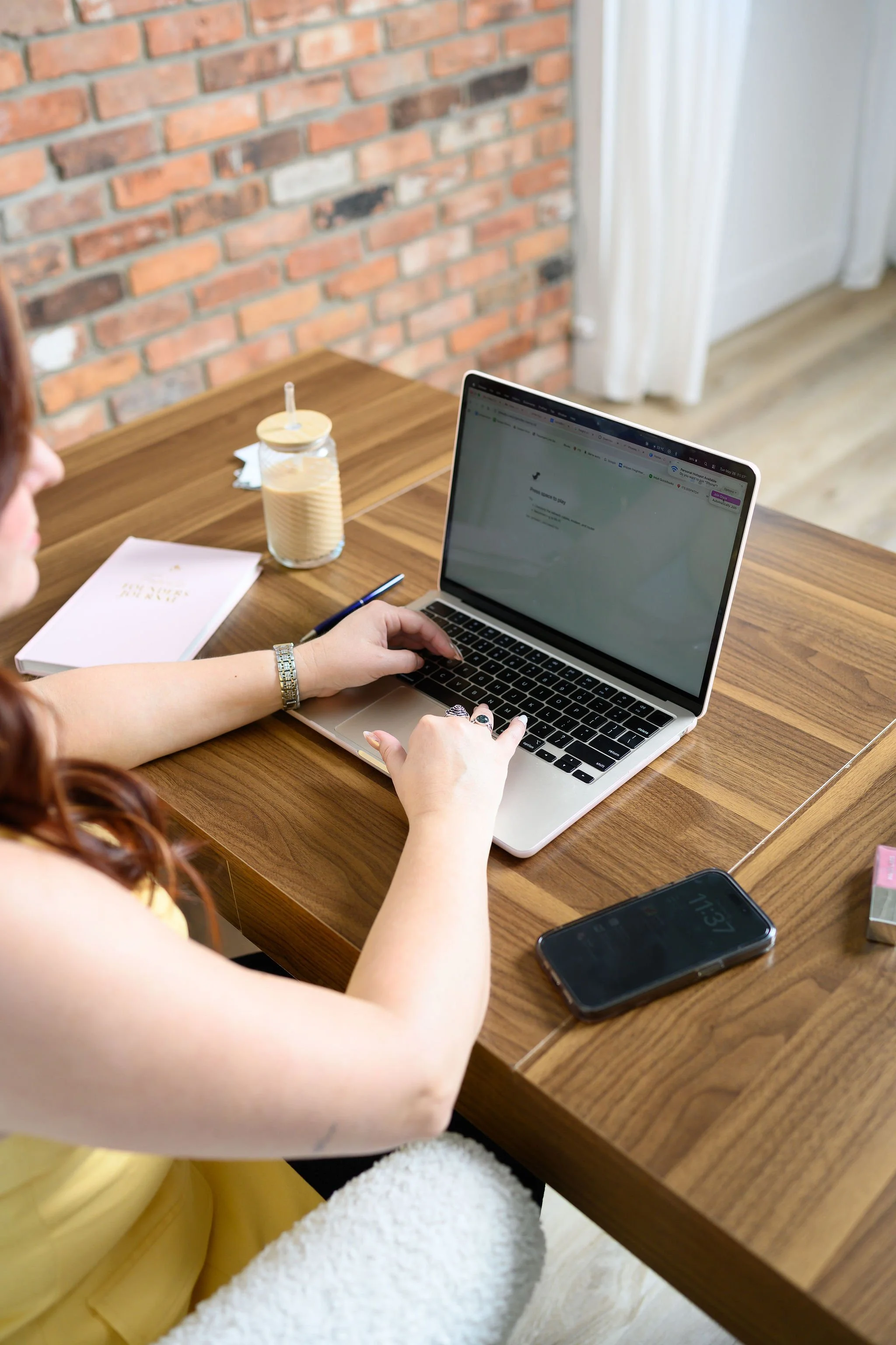 A person wearing a yellow dress with a white fluffy skirt working at a wooden table with a silver laptop, a notebook, pen, two glasses of iced coffee, and a black smartphone.