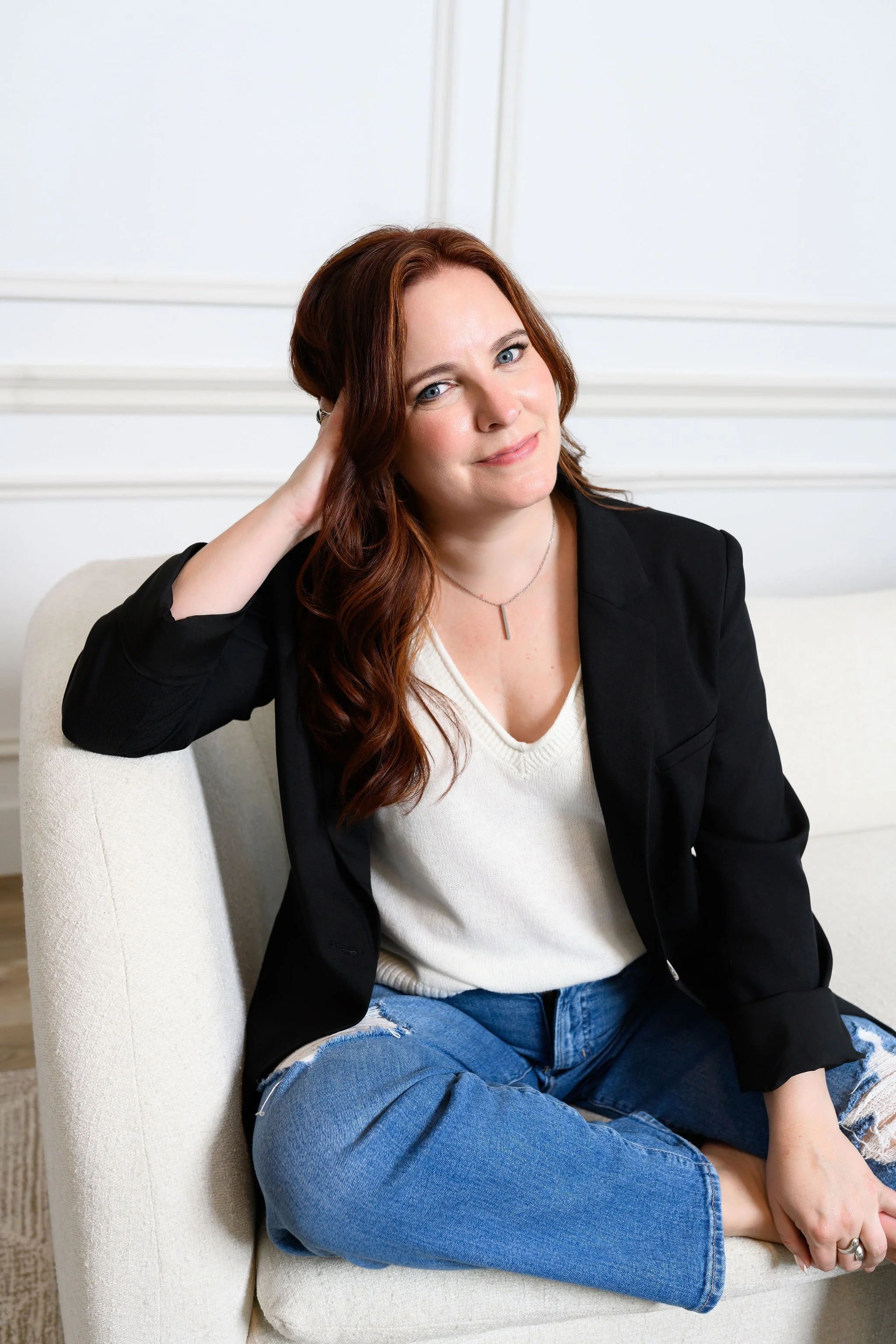 A woman with long, wavy auburn hair and blue eyes sitting on a beige chair against a white wall. She is wearing a black blazer over a white top, ripped jeans, and a simple silver necklace. She has a gentle smile and her right hand resting on her neck.