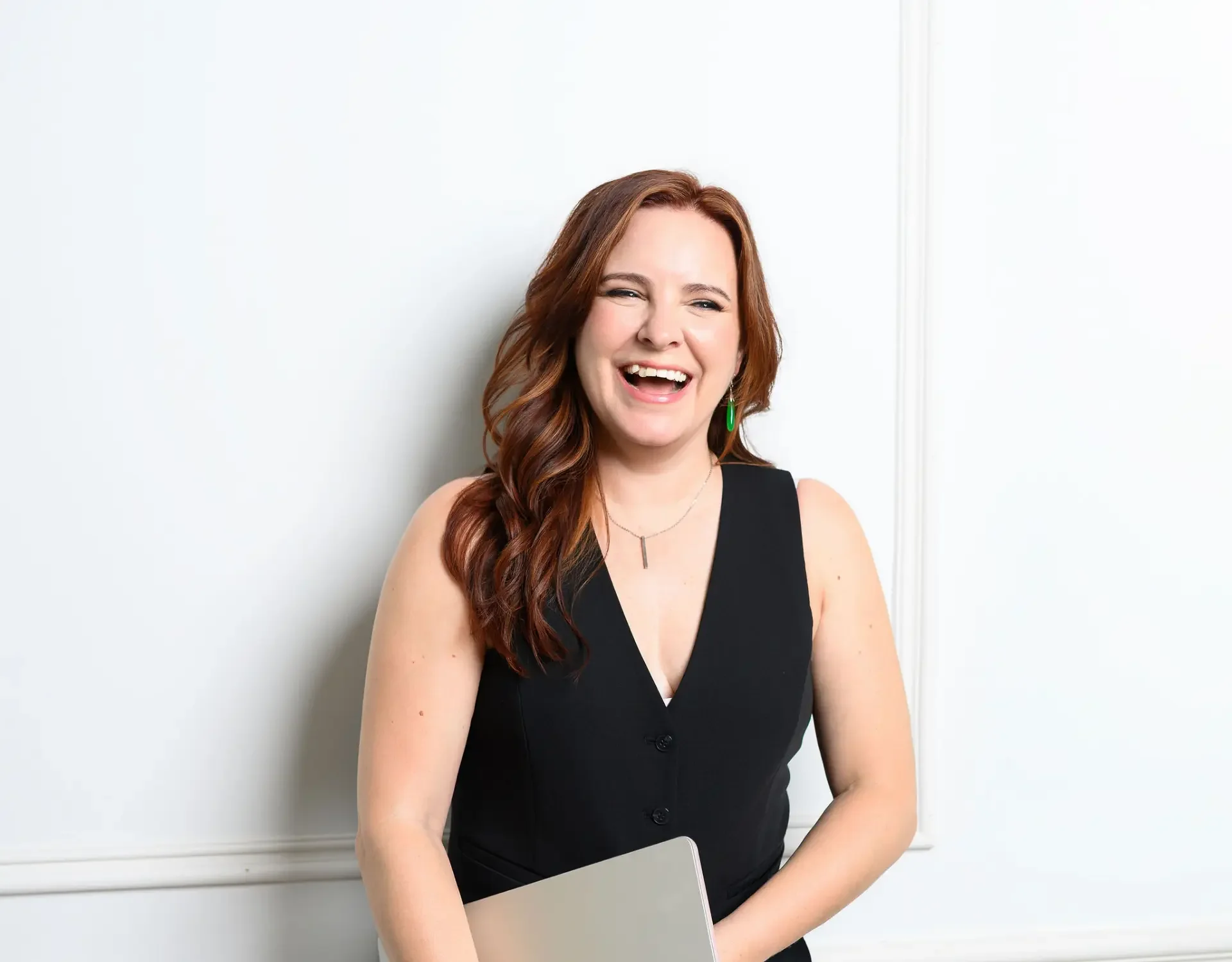 A woman with red hair wearing a black sleeveless dress laughing, standing against a white wall, holding a laptop.
