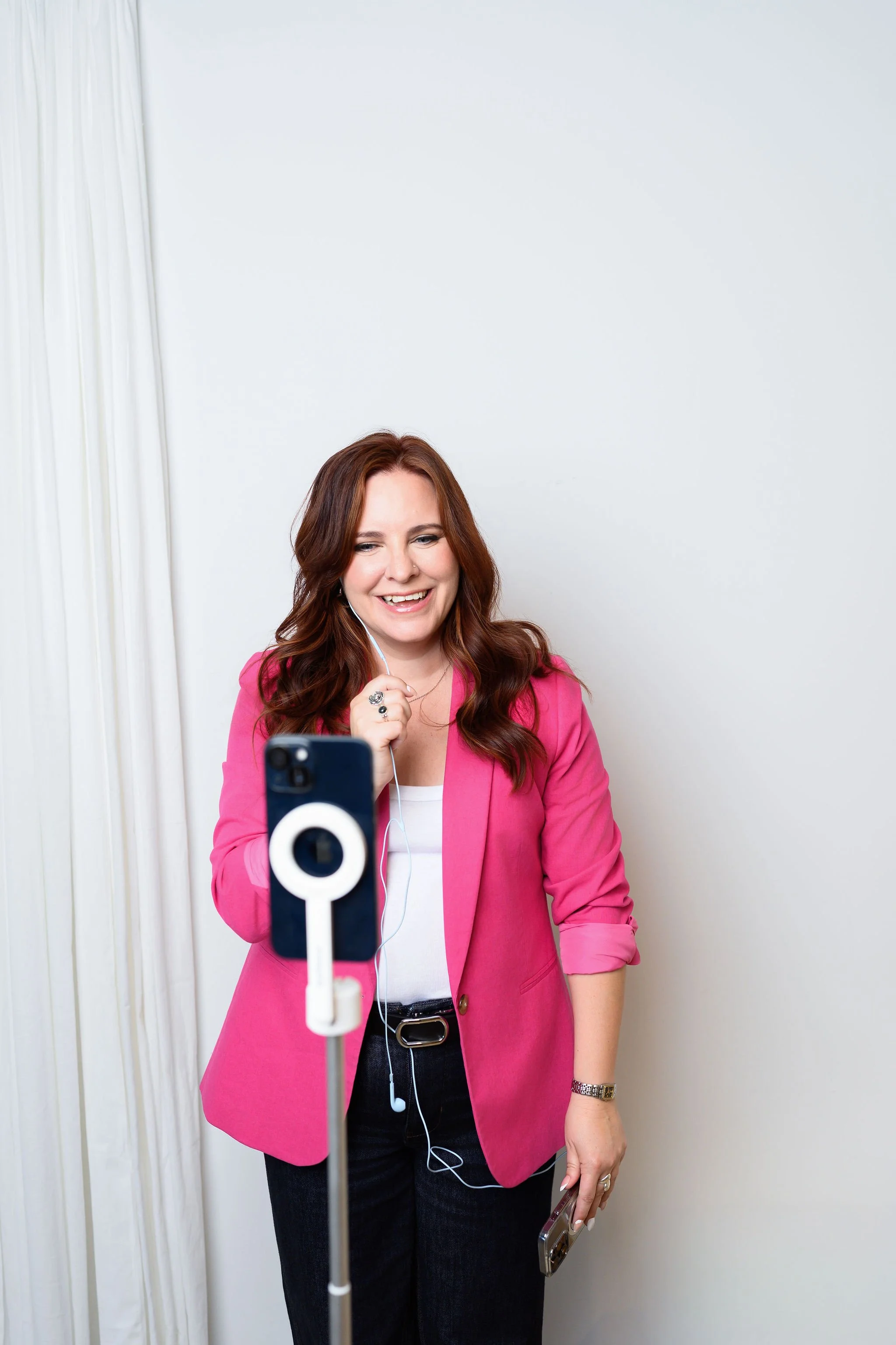 A woman with long red hair wearing a pink blazer, white top, and dark jeans, smiling as she records a video call on her smartphone, with white earbuds in her ears, standing against a plain white wall.