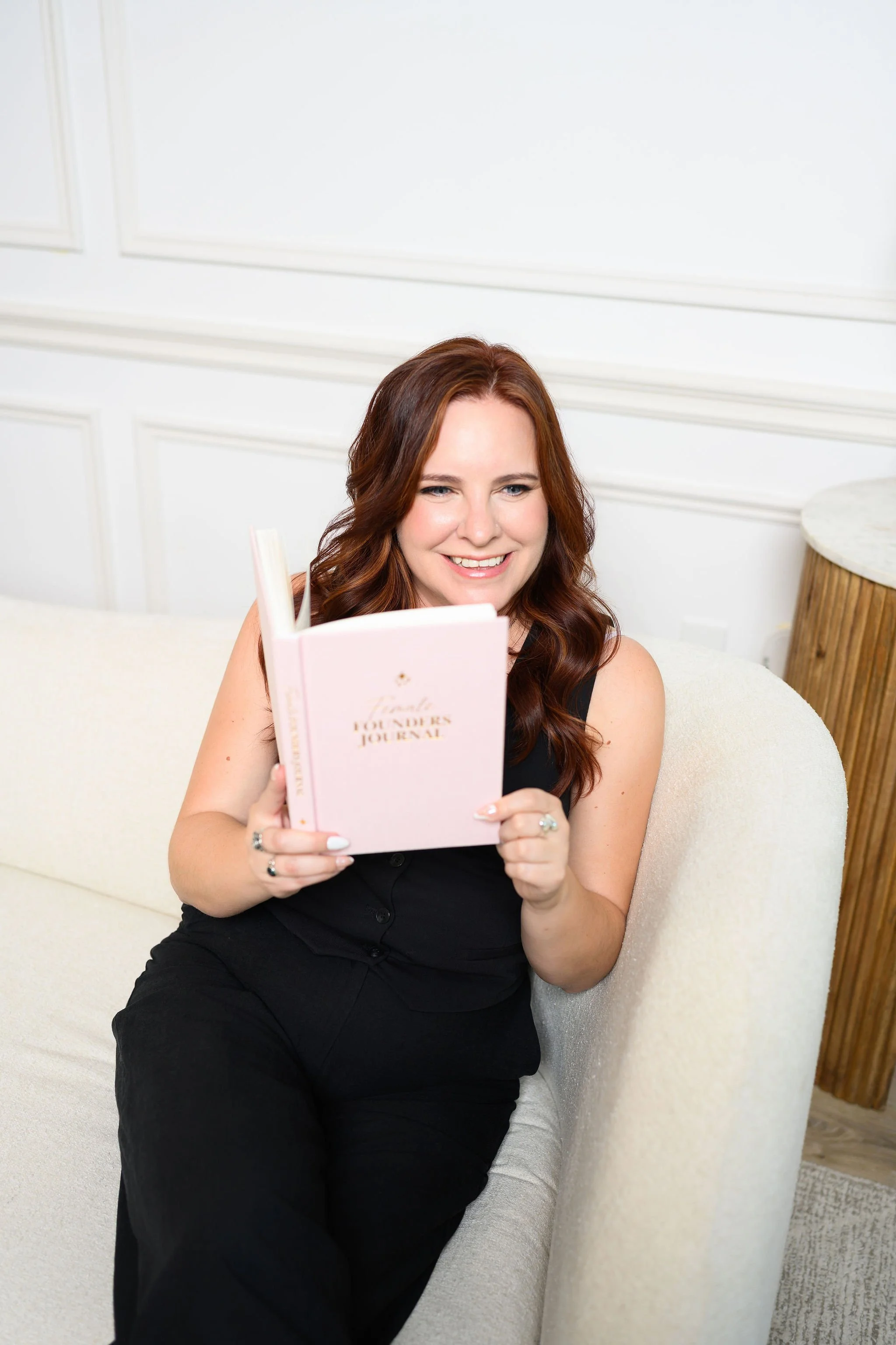 A woman with long, wavy red hair sitting on a cream-colored sofa, smiling and holding a pink book titled 'Lindie' with the subtitle 'Founders Journal' inside.