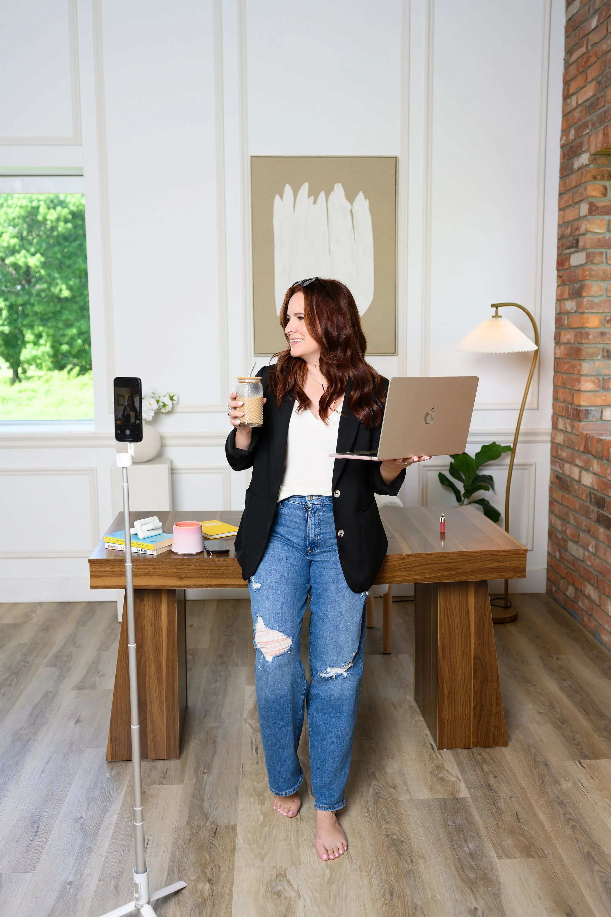 Woman with wavy brown hair, wearing a black blazer and ripped jeans, holding a glass jar with a straw, standing barefoot in a bright room with a large window, a wooden table, and a brick wall.