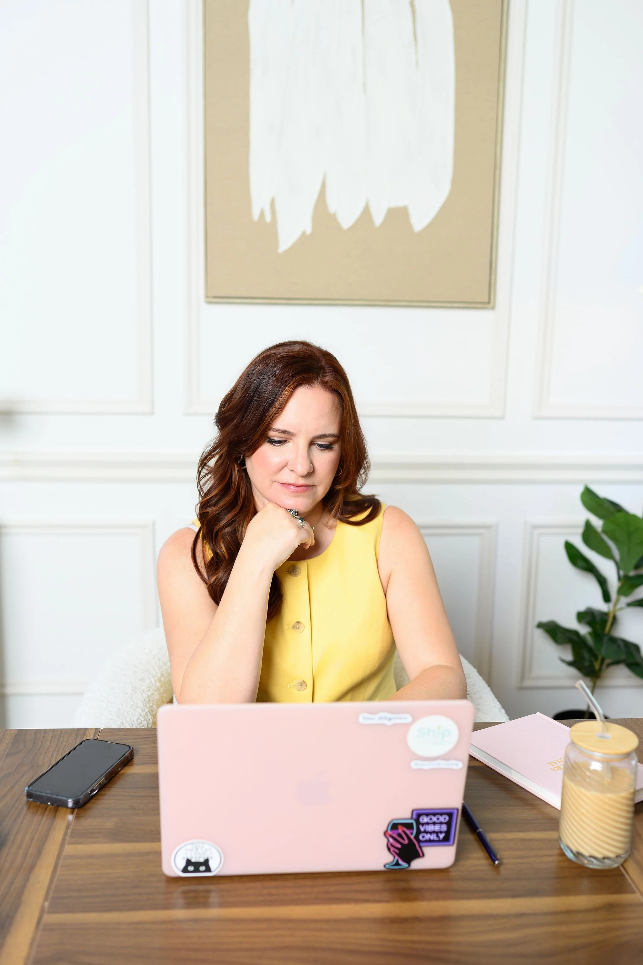 A woman with red hair wearing a yellow sleeveless top sitting at a wooden table, working on a pink laptop decorated with various stickers. The background includes a framed piece of artwork, a white wall with paneling, a green plant, a smartphone, a notebook, and a glass jar on the table.