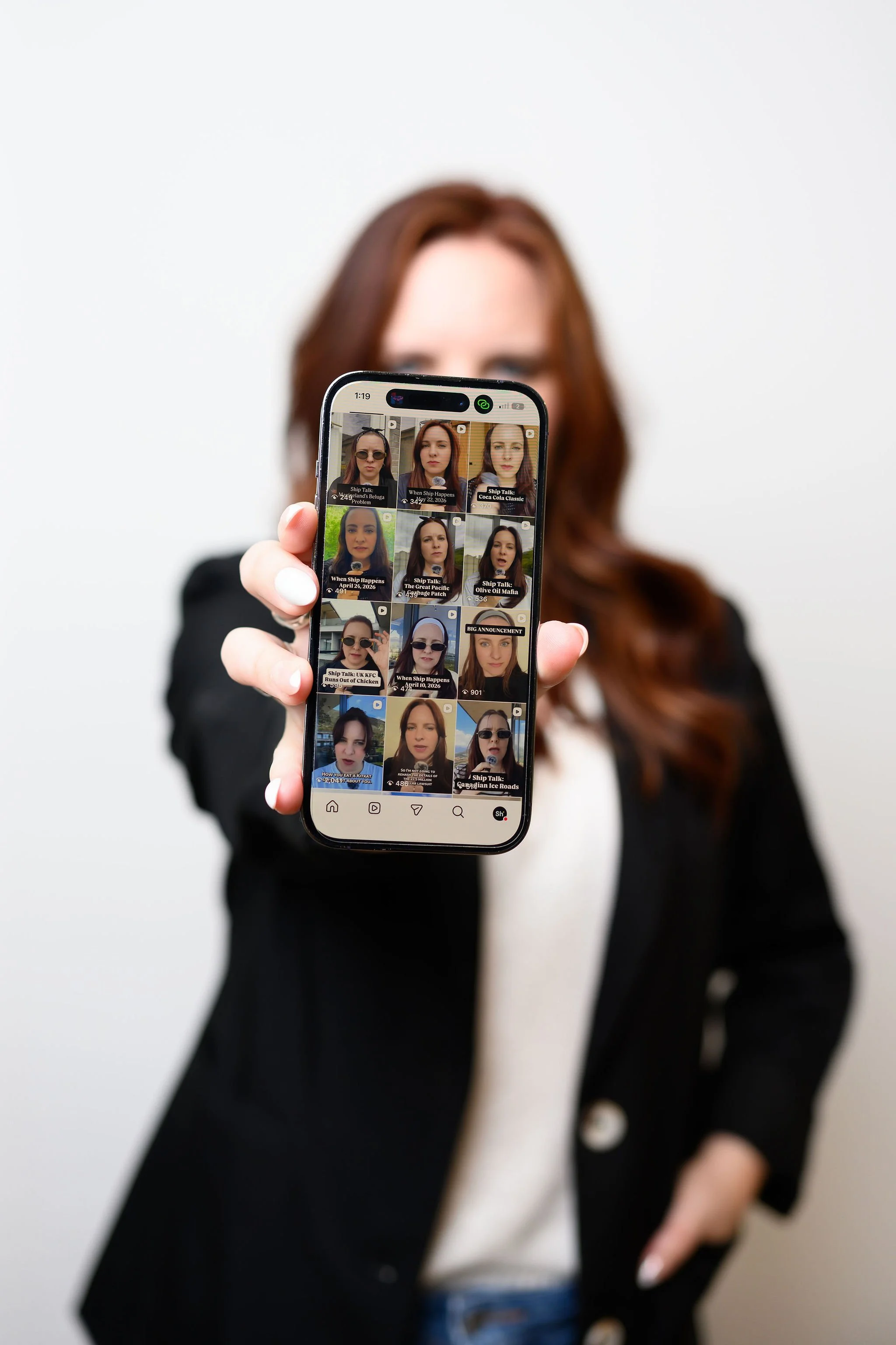 A person with long brown hair holding a smartphone showing a grid of selfies and video thumbnails, with a plain white background.