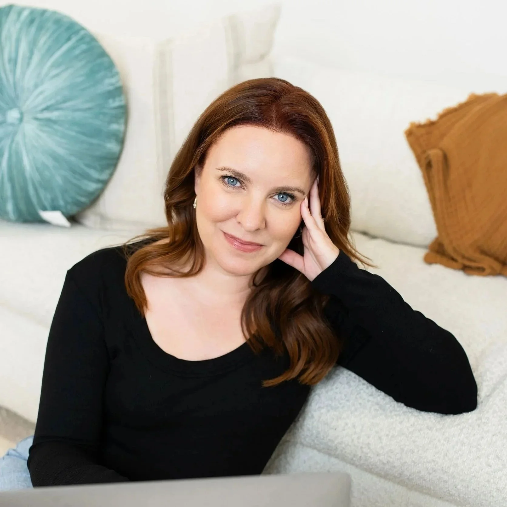 Jennifer with red hair and blue eyes leaning on a beige couch, resting her head on her hand, looking at the camera with a slight smile.