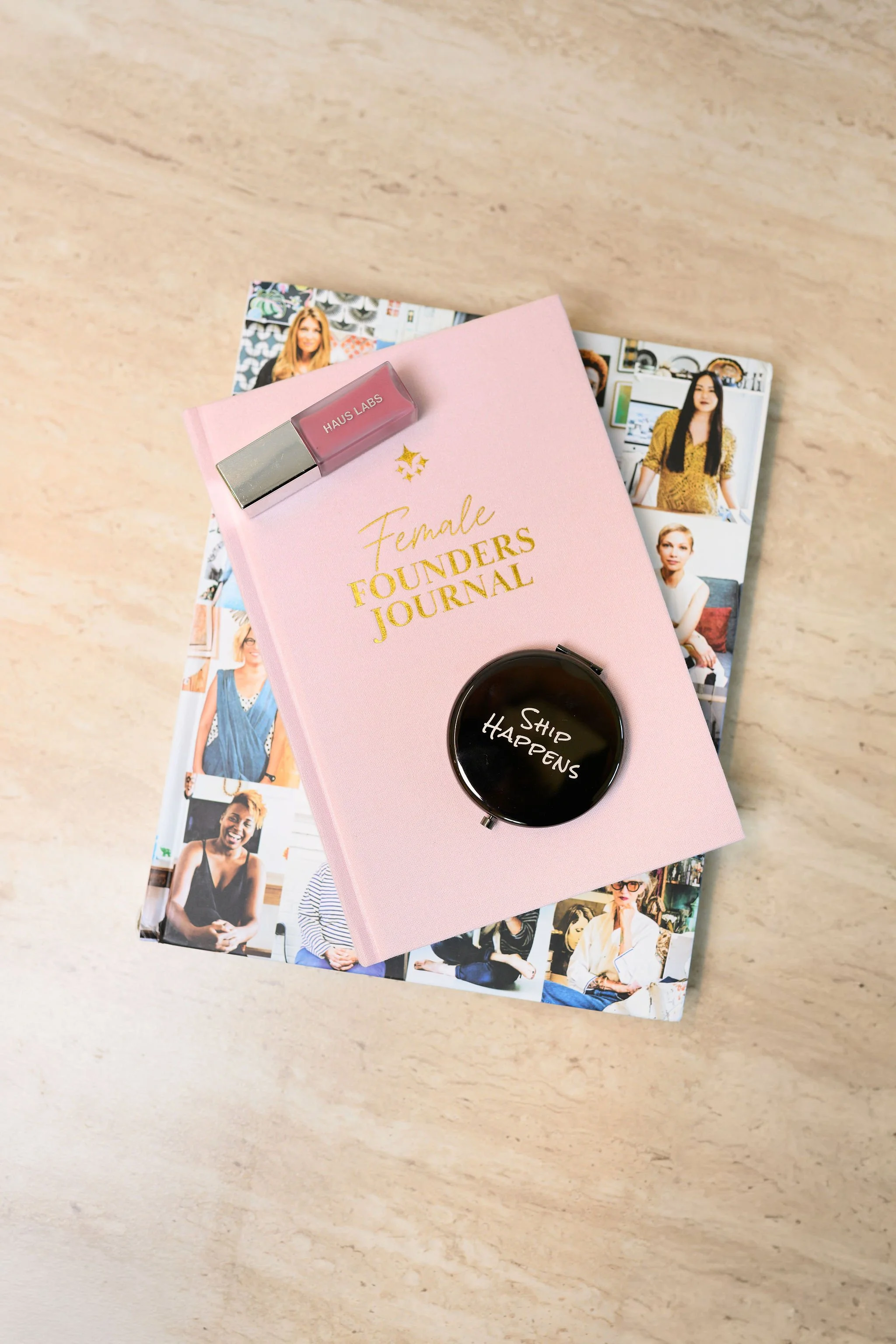 A pink notebook labeled 'Female Founders Journal' with a small pink and silver Haus Labs lipstick on top, a black mug with 'Ship Happens' written on it, and a magazine with various photos of women on a light-colored surface.