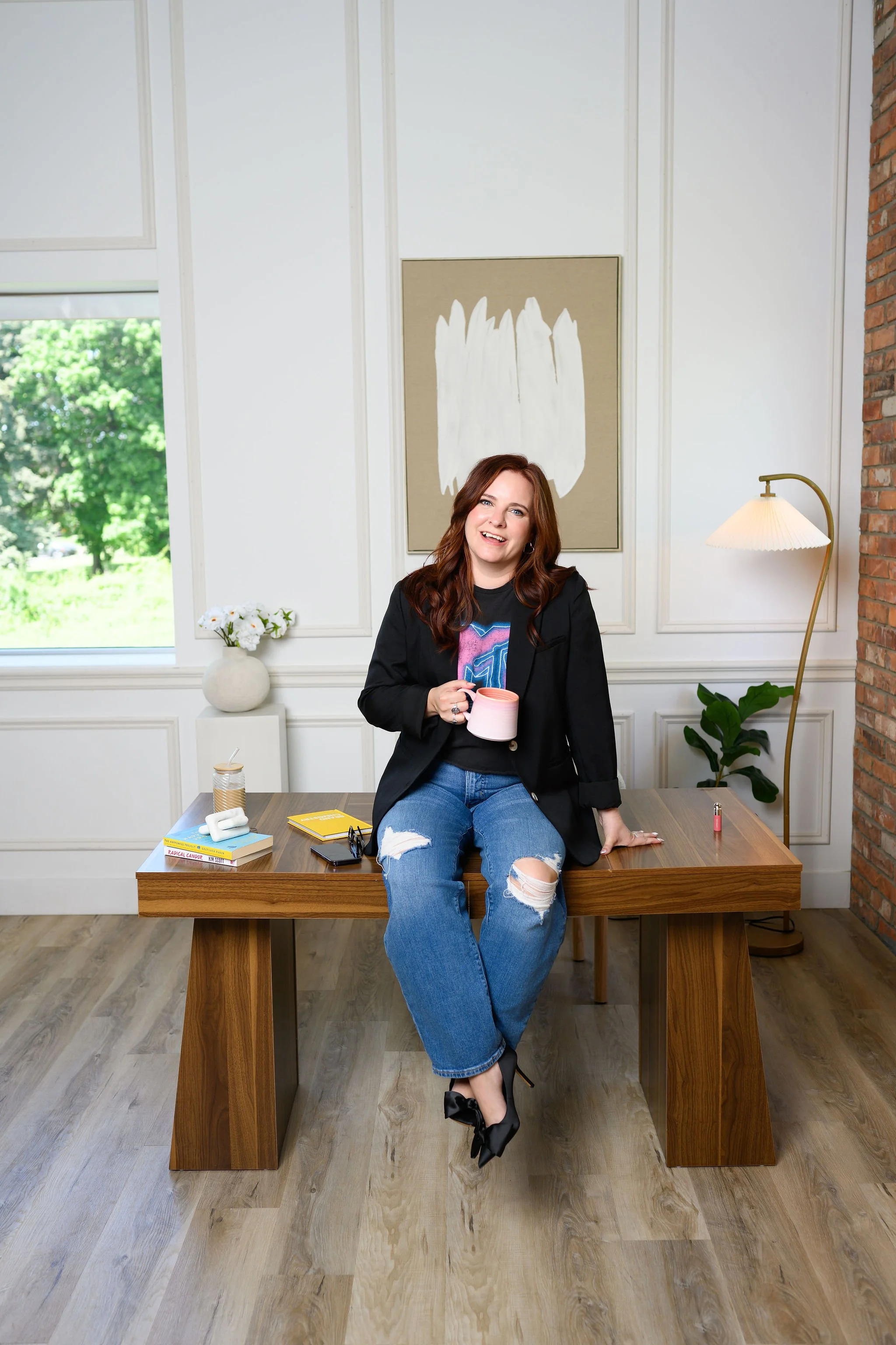 A woman with long reddish-brown hair wearing a black jacket and ripped jeans sitting on a wooden table, holding a pink mug, smiling. The room has white walls, a large window with green trees outside, a modern floor lamp, a houseplant, books, and decorative items on the table.