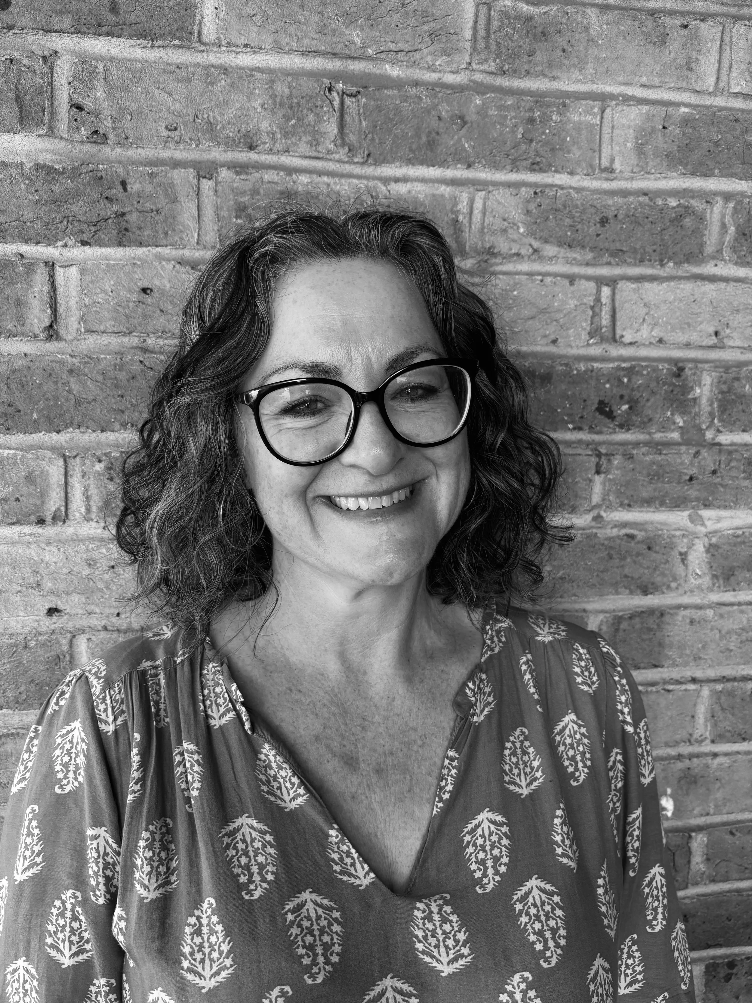 A woman with curly hair and glasses smiling, standing against a brick wall, in black and white.