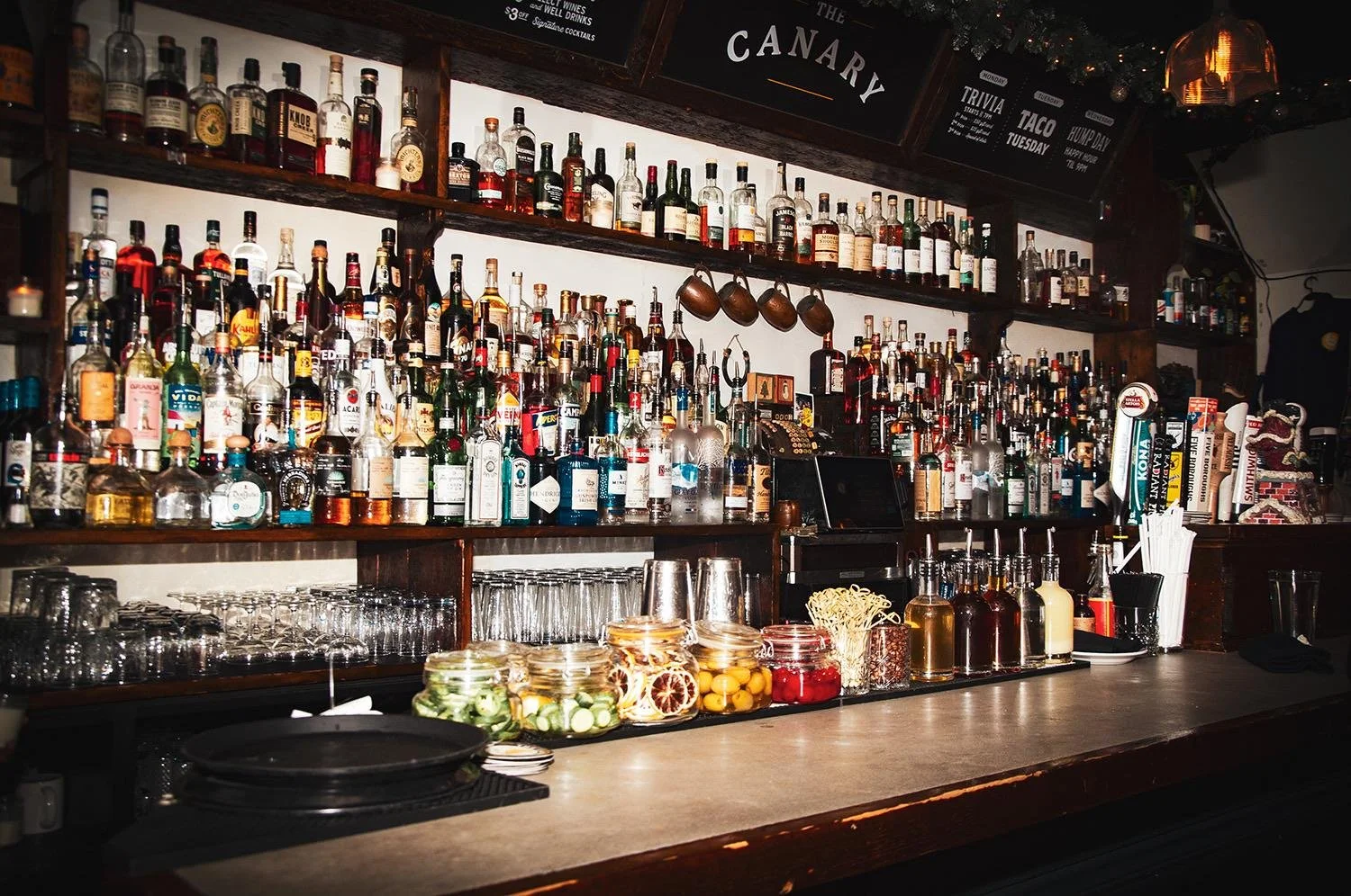 Bar counter with numerous bottles of liquor, glassware, and garnishes, with a dark wooden shelf background and dim lighting.