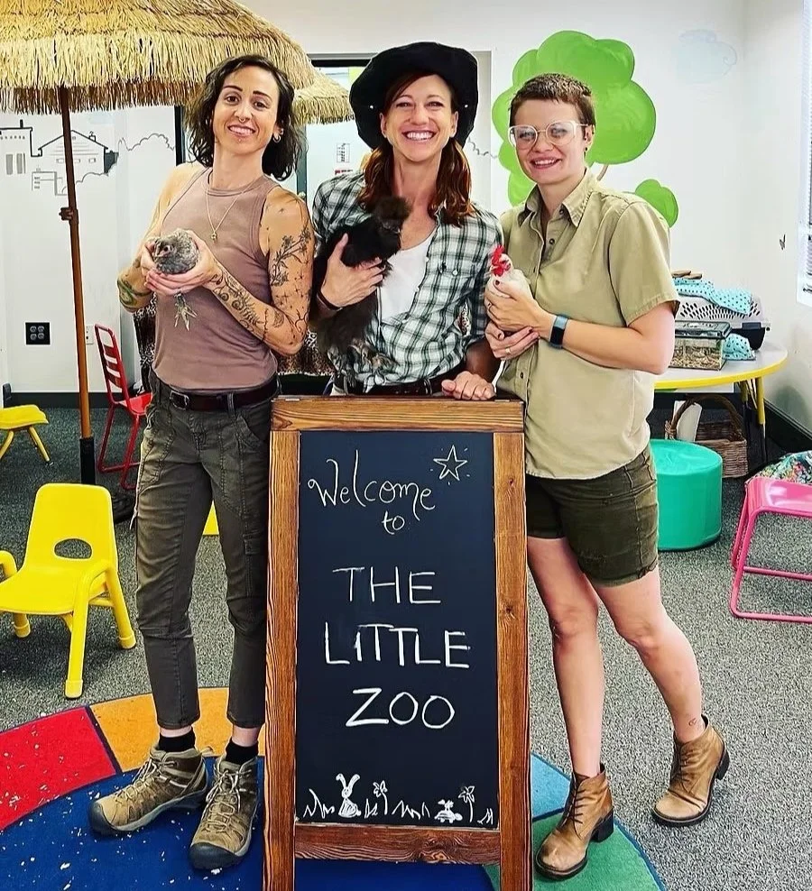 Three women standing behind a chalkboard sign that says 'Welcome to The Little Zoo,' each holding a small animal, in a children’s playroom decorated with a thatched roof and tree illustration.
