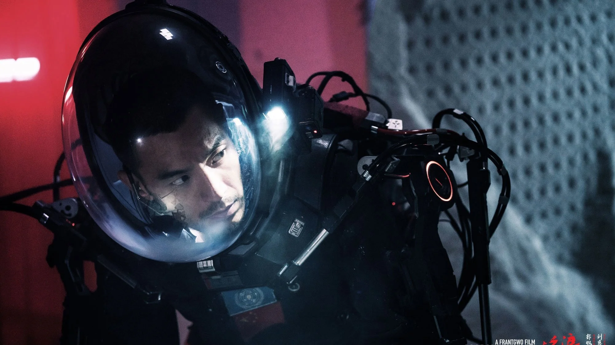 A man in a space suit with a helmet, connected to various wires and equipment, inside a spacecraft or space station.