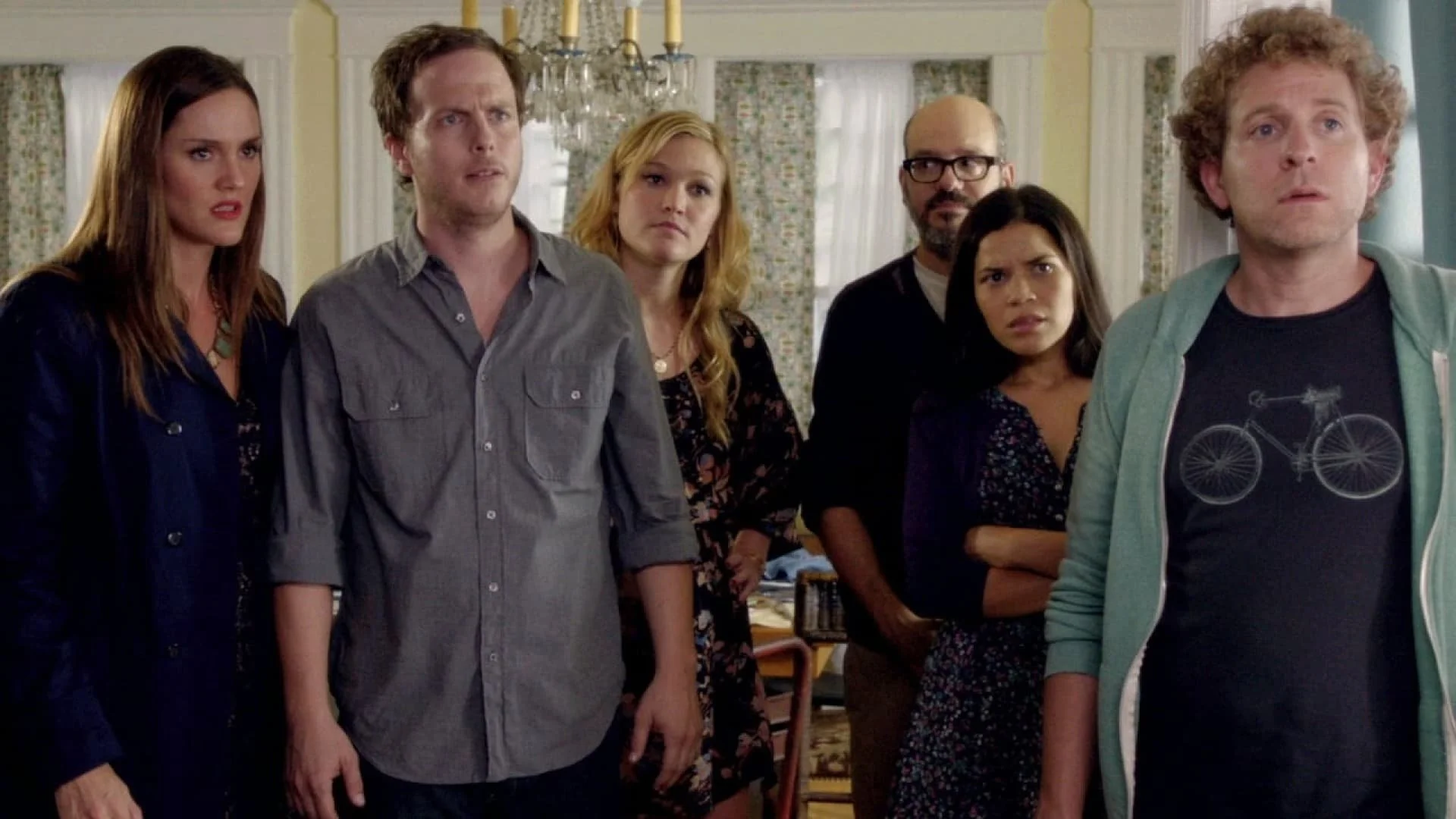 Six people standing inside a room, looking towards the right with serious and confused expressions.