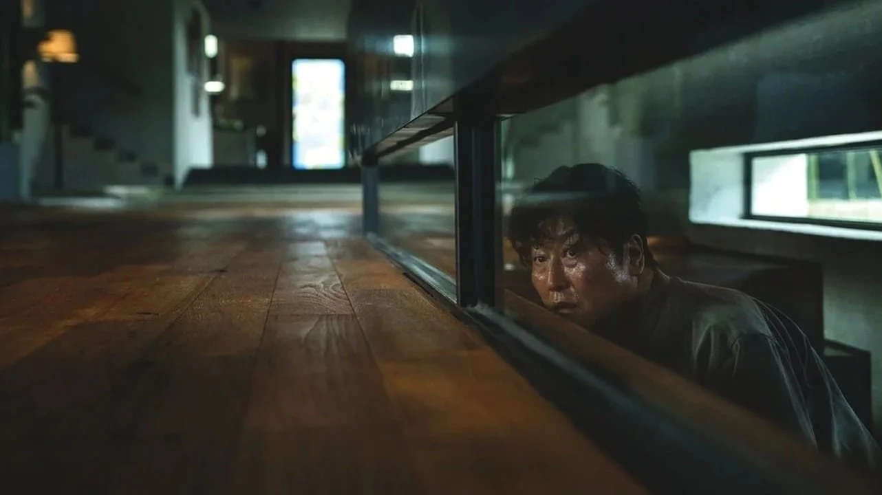 A person peering underneath a table through a glass panel, with a wooden floor and a bright door in the background.