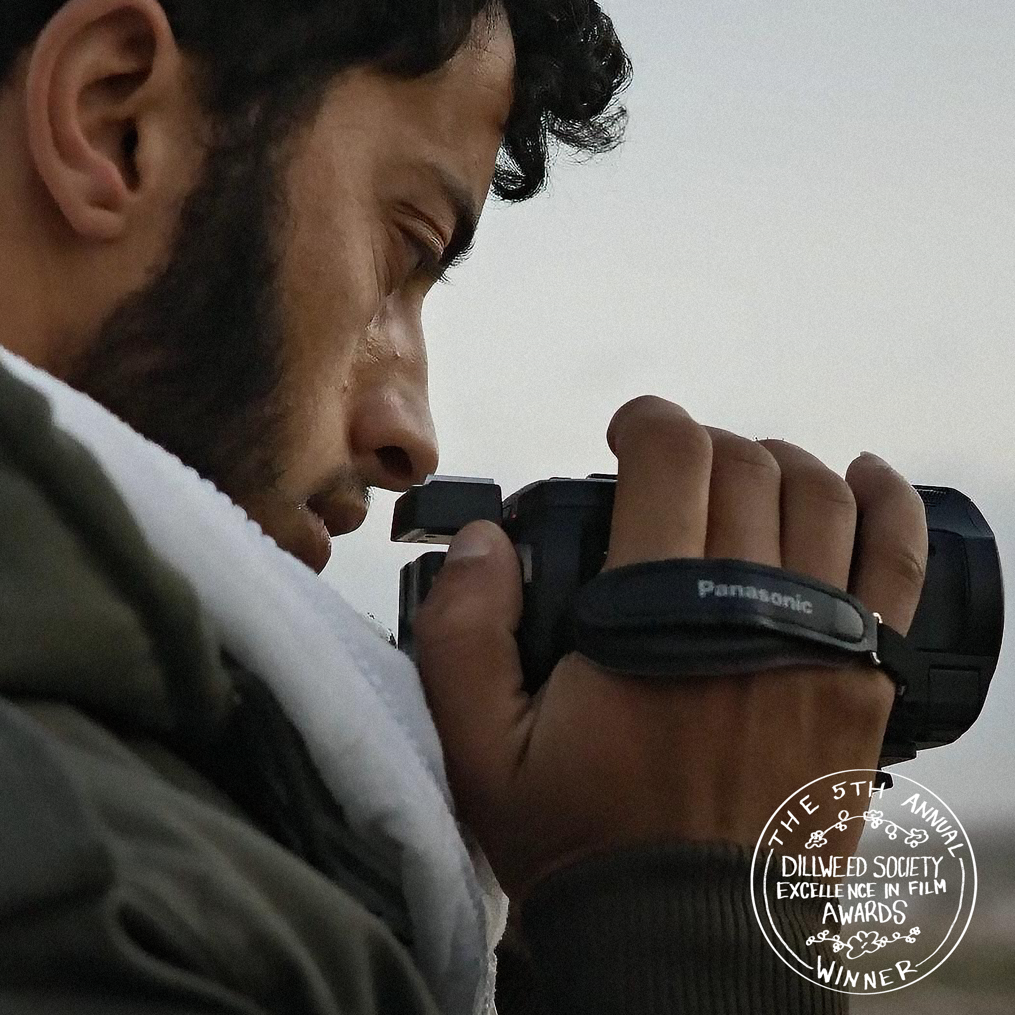 A man with dark, curly hair and a beard holding a Panasonic camera close to his face, looking down while taking a photo. He is wearing a green jacket and a white shirt or hoodie underneath. There is a circular emblem in the bottom right corner that says 'The 5th Annual Dillweed Society Excellence in Film Awards Winner.'