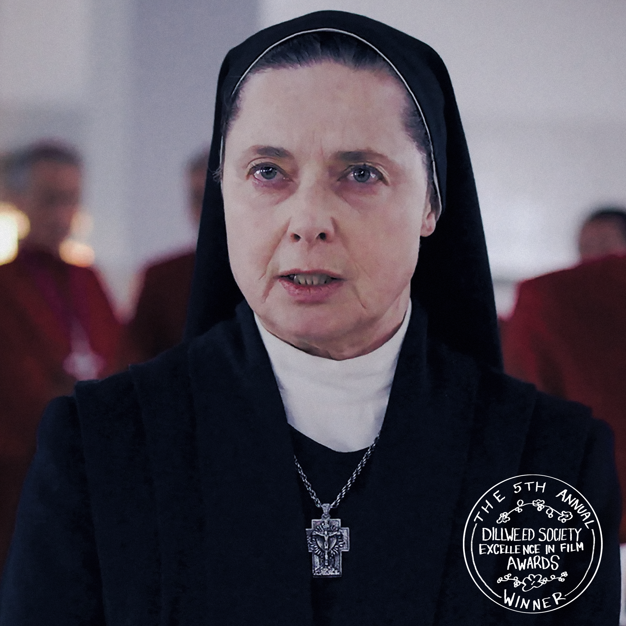 A nun with a stern expression, wearing a black habit and a white clerical collar, with a cross necklace, standing in a room with other nuns in the background.