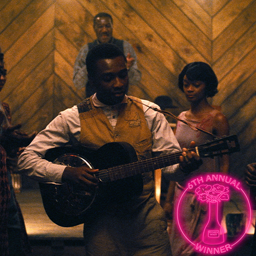 A man playing tennis outdoors with trees in the background, holding a red tennis racket, wearing a sleeveless athletic shirt, and a circular award badge in the bottom right corner indicating he is a winner of the 5th Annual Dillweed Society Excellence in Film Awards.