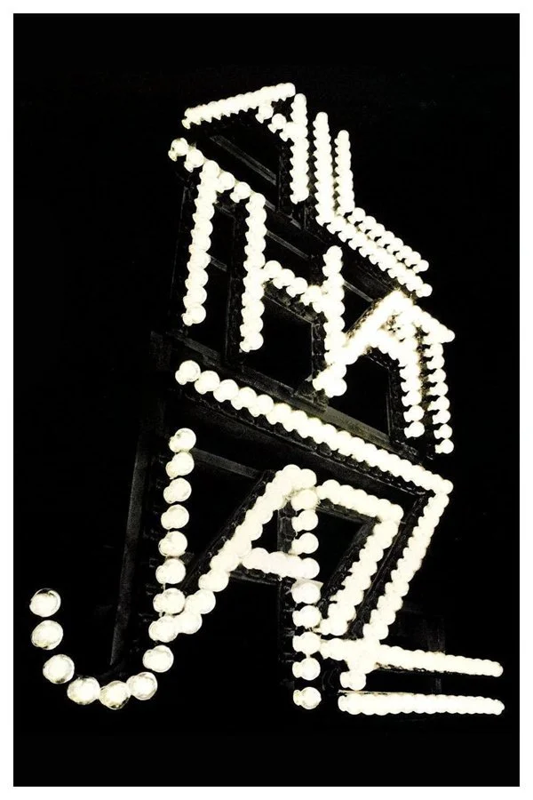A zigzagging neon sign composed of white round bulbs, creating a stylized arrow pointing to the right on a black background.