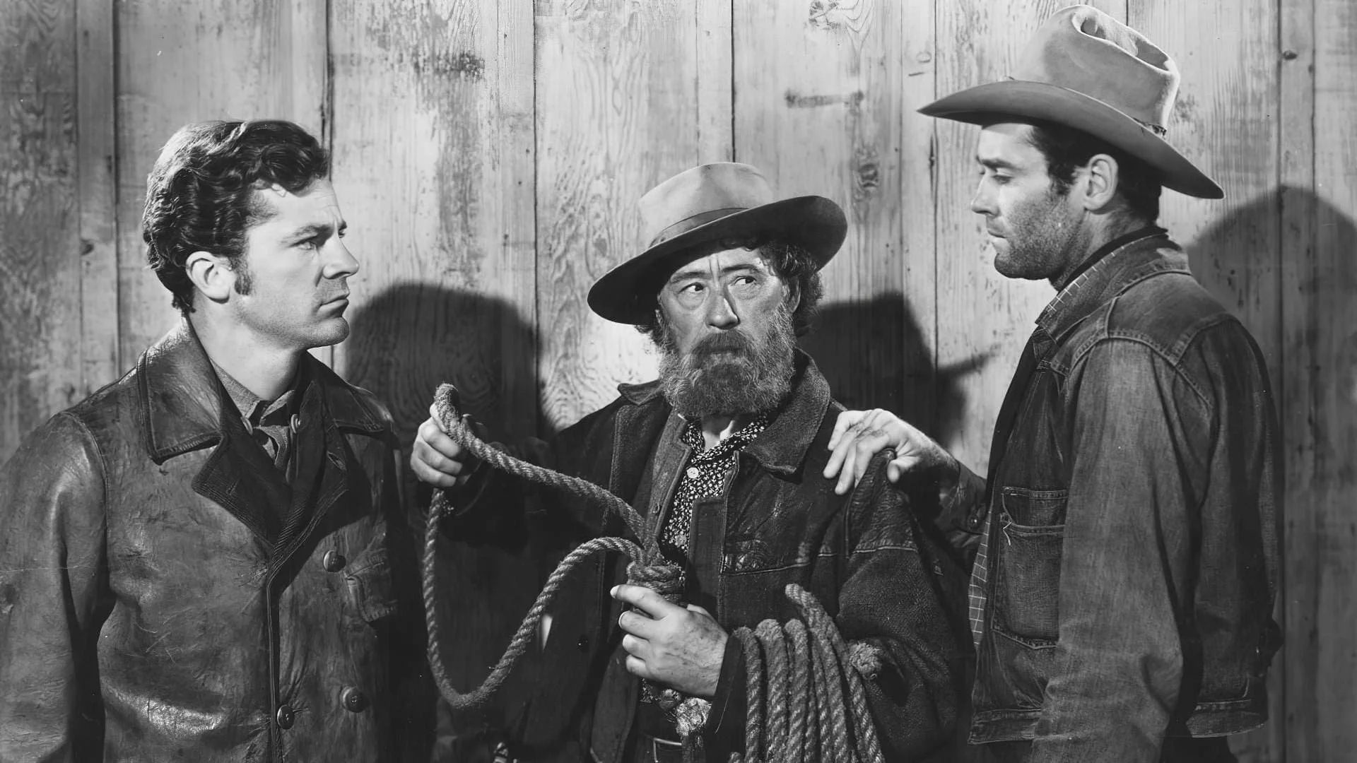 Three men in cowboy attire face each other indoors, with a wooden wall in the background. The man in the middle has a beard, wears a hat, and is holding a noose. The two men on either side are looking at him; one has short dark hair and the other has a hat and longer hair.