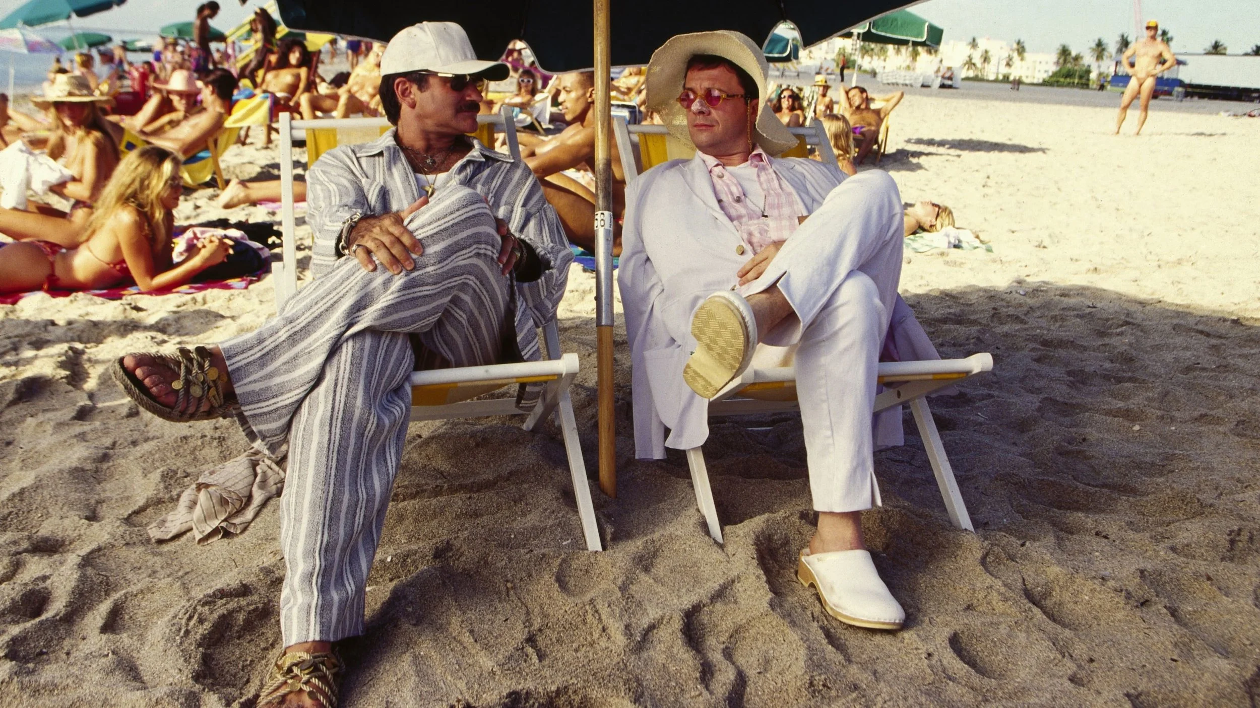 Two men in vintage beach attire sitting under a large umbrella, talking, with many beachgoers in the background.