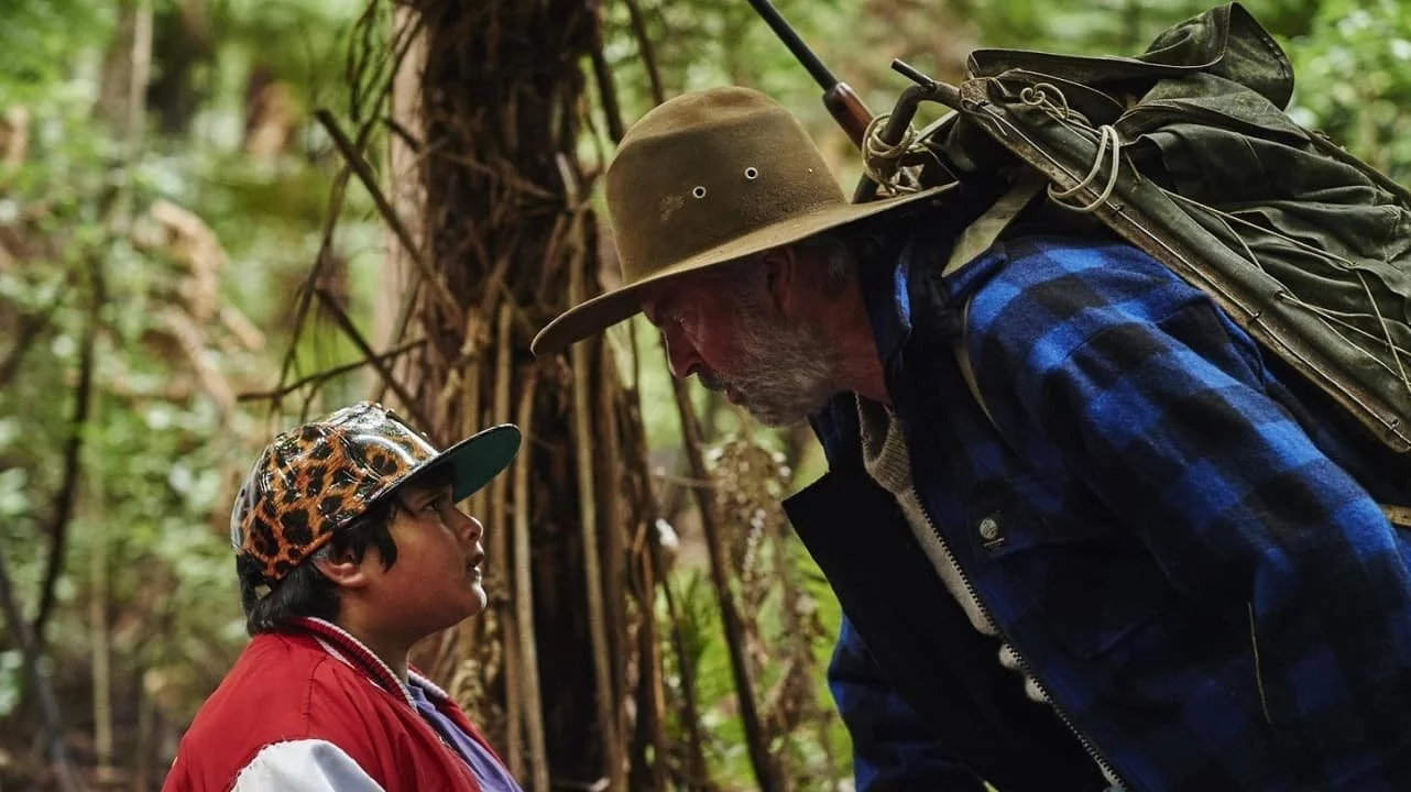 An older man and a young boy in a forest, facing each other, with the man leaning down to the boy. The man is wearing a wide-brimmed hat, a blue plaid jacket, and carrying a backpack. The boy is wearing a leopard-print baseball cap and a red jacket.