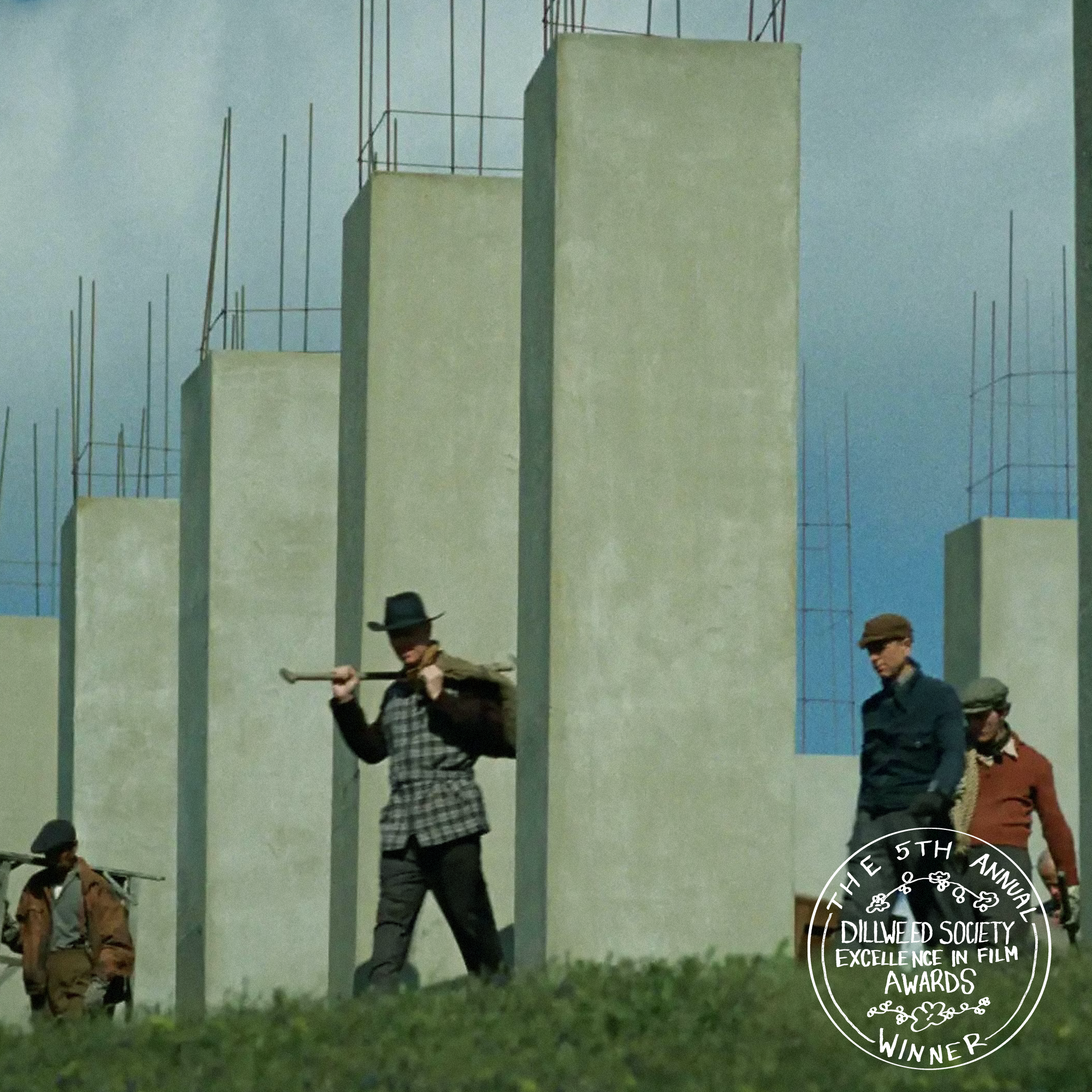 Three men dressed in vintage clothing walking among concrete columns at a film set with a cloudy sky in the background, featuring a circular badge in the bottom right corner that reads 'The 5th Annual Dillweed Society Excellence in Film Awards Winner'.