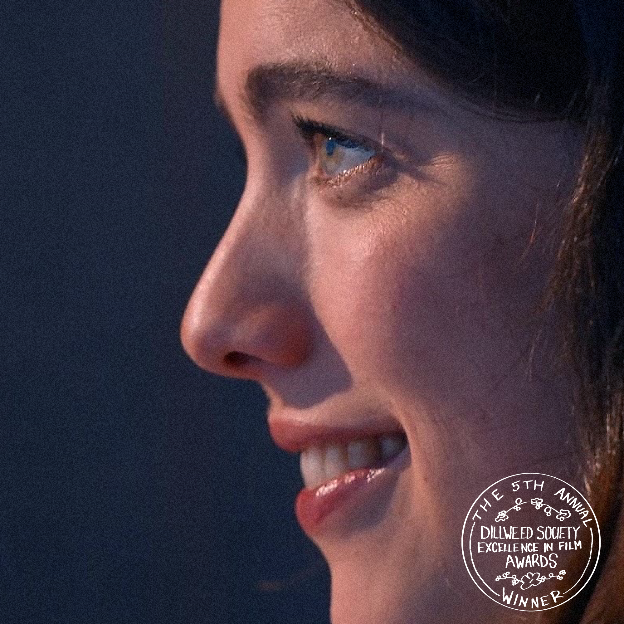 Close-up of a woman's face in profile, smiling with visible teeth, showing brown eyes, smooth skin, and dark hair, with a dark background and an award emblem in the bottom right corner.