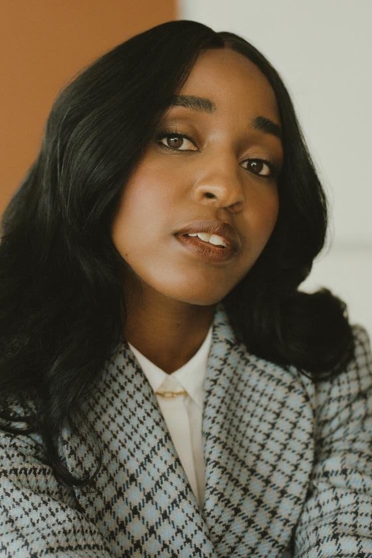 A woman with long black hair wears a checkered blazer and white collared shirt, looking at the camera with a neutral expression. DILLIES