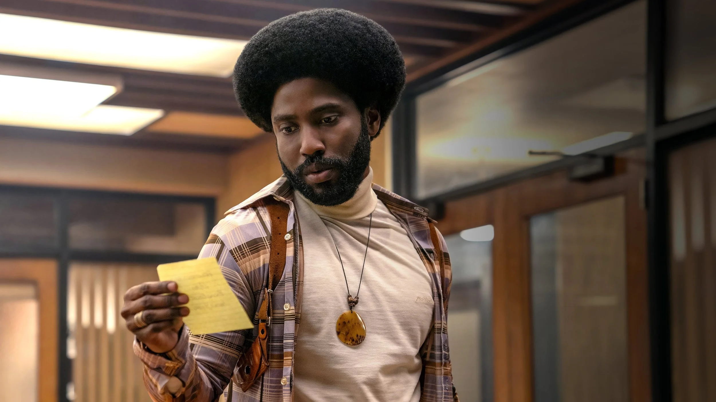 A man with a dark skin tone, full beard, and large afro hairstyle holds a yellow sticky note in a room with wood-paneled walls and large windows.
